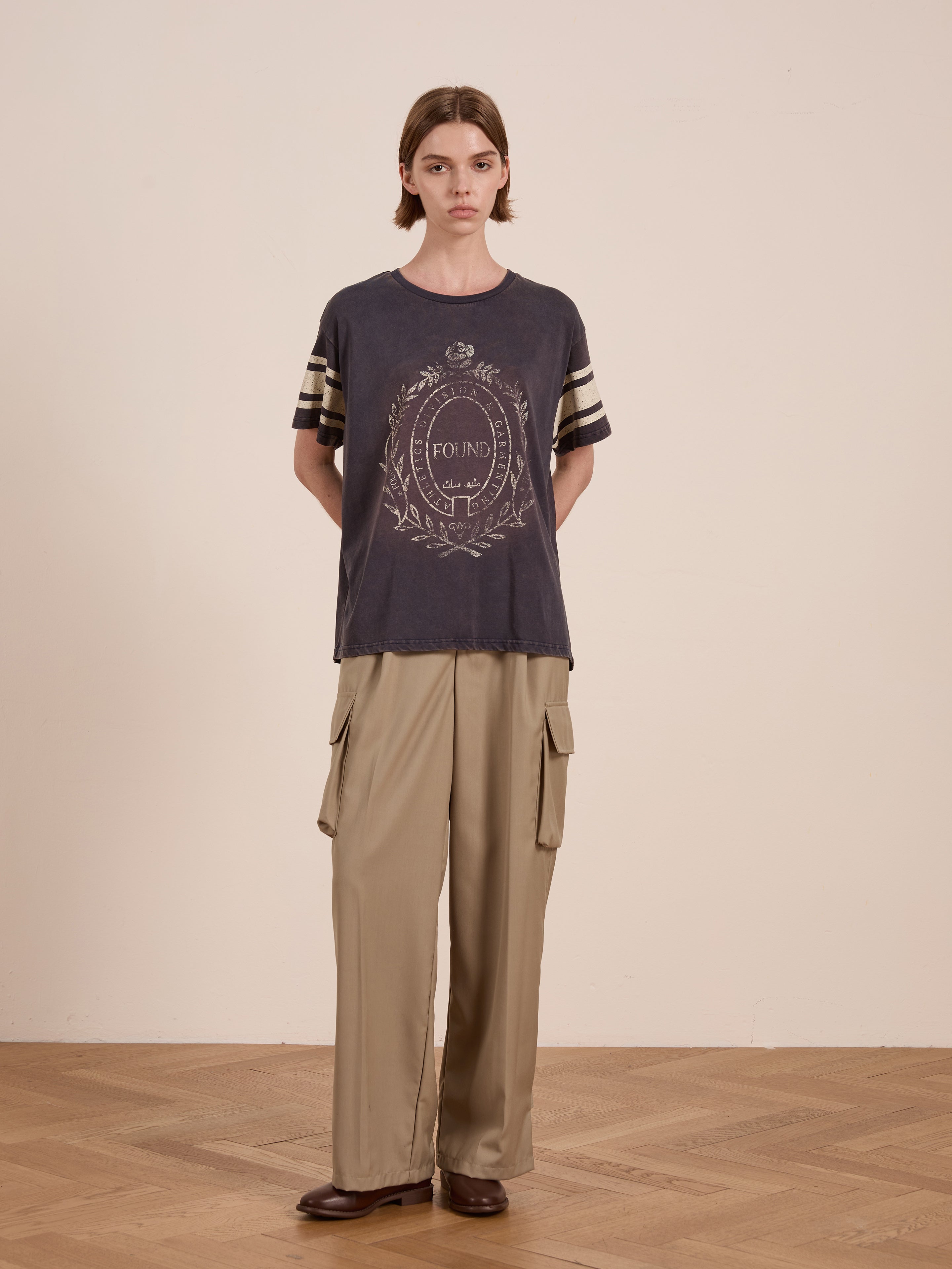 A person in a FOUND Collegiate Crest Tee with a unisex relaxed fit and khaki cargo pants stands on a wooden floor against a plain wall.