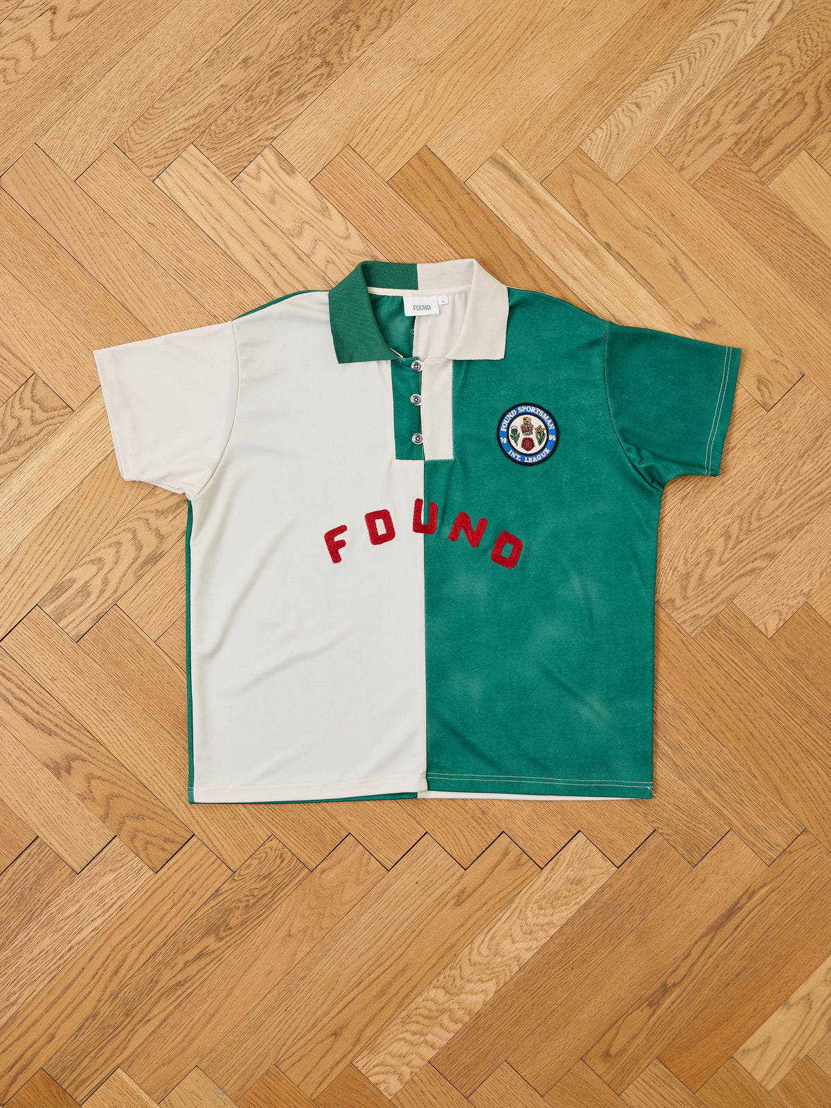 The Color Block Regent Jersey by FOUND is a short-sleeve polo shirt featuring a half white, half green color block design, bold red “FOUND” text, and a circular patch on the left chest. It is displayed laid flat on a wooden floor for a classic vintage sportswear-inspired look.