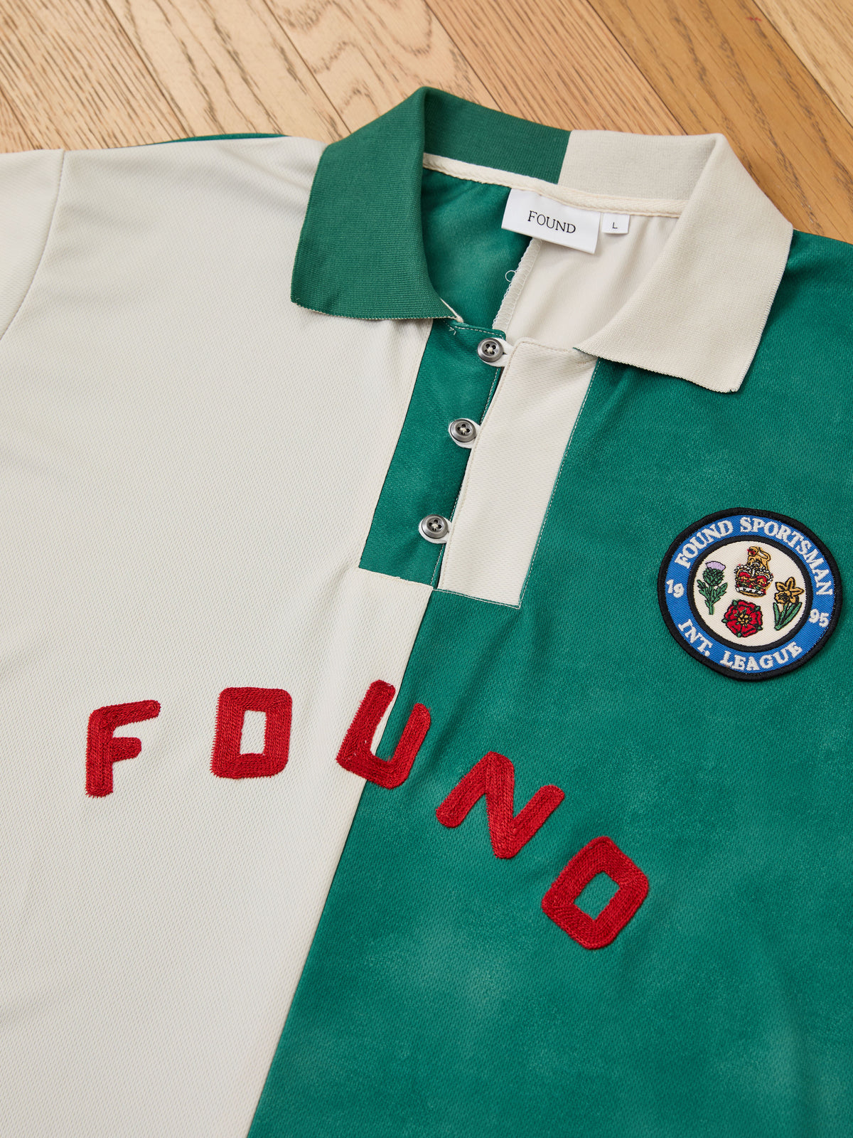 A vintage sportswear-inspired Color Block Regent Jersey by FOUND, featuring green and cream color blocking, a collared design, “FOUND” in red letters, and a circular chest patch reading “FOUND SPORTSMAN ART. LEAGUE,” laid flat on a wooden floor.