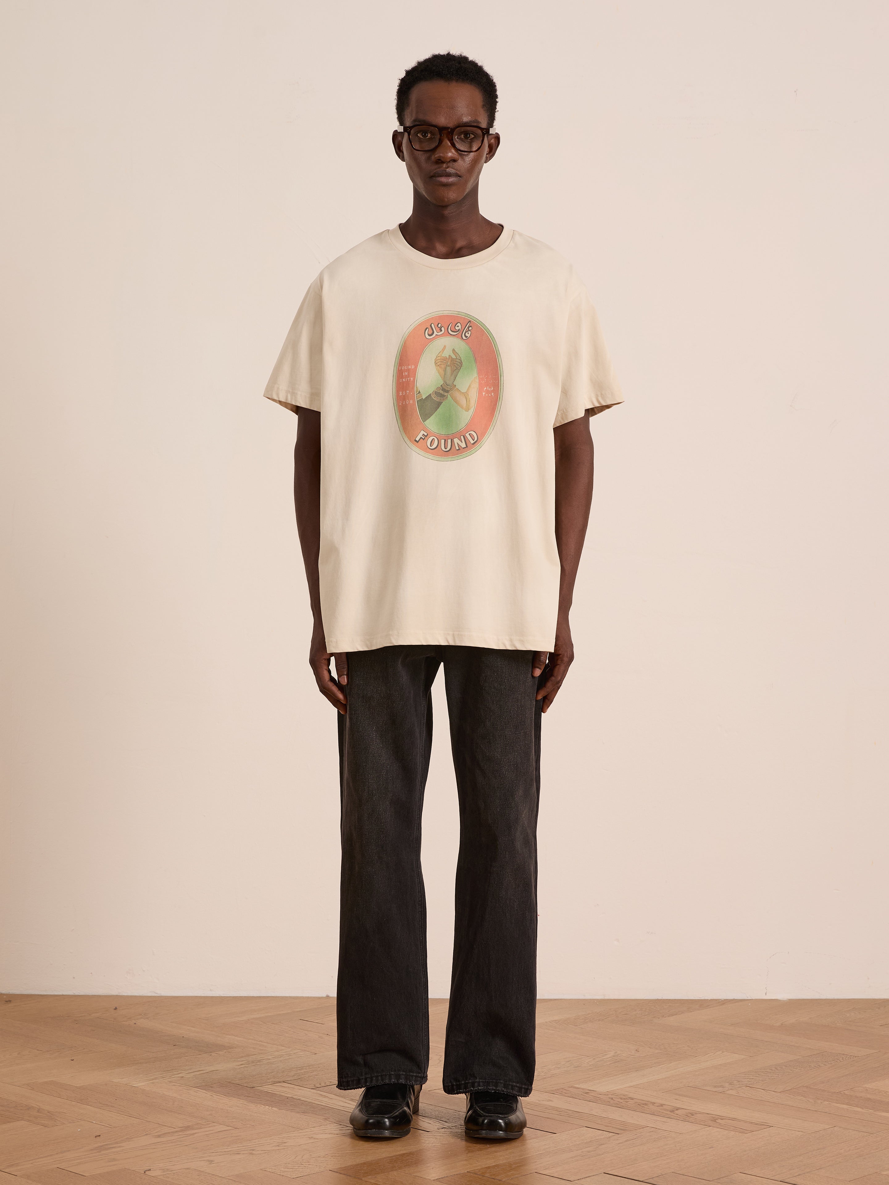 Indoors, someone wears glasses and the FOUND Community Hands Tee—a light-colored, oversized unisex cotton shirt with vintage matchbox artwork and "FOUND IN UNITY" text—paired with dark jeans and black shoes against a plain backdrop.