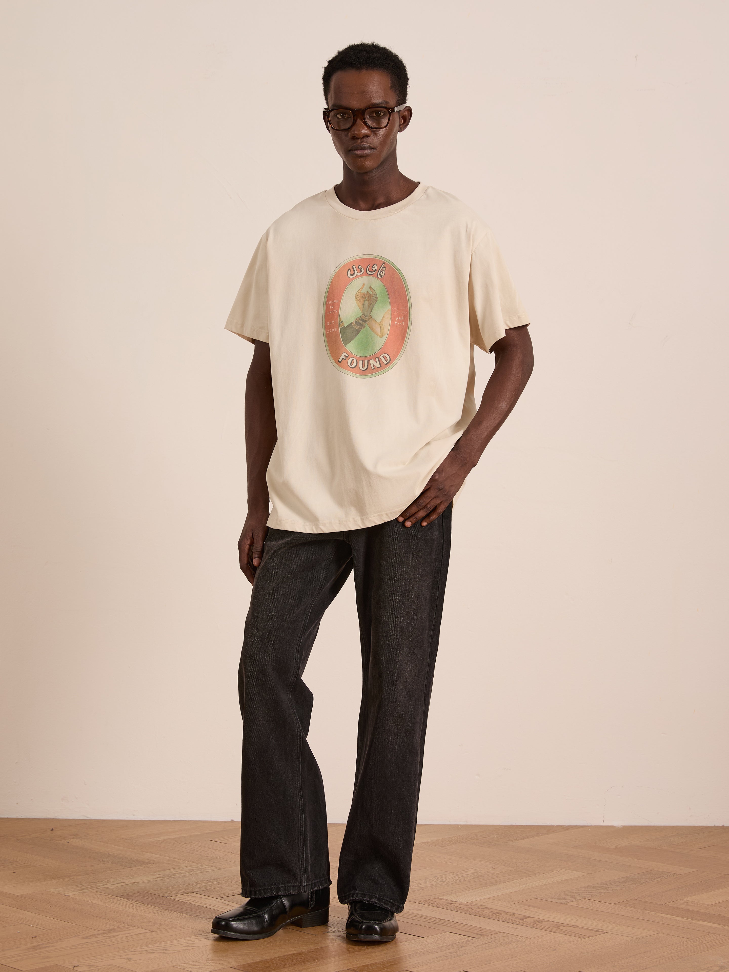 Indoors, a person models the FOUND Community Hands Tee—a cream unisex cotton shirt with vintage matchbox art—paired with dark jeans, black shoes, and glasses, against a plain light wall and wooden floor.