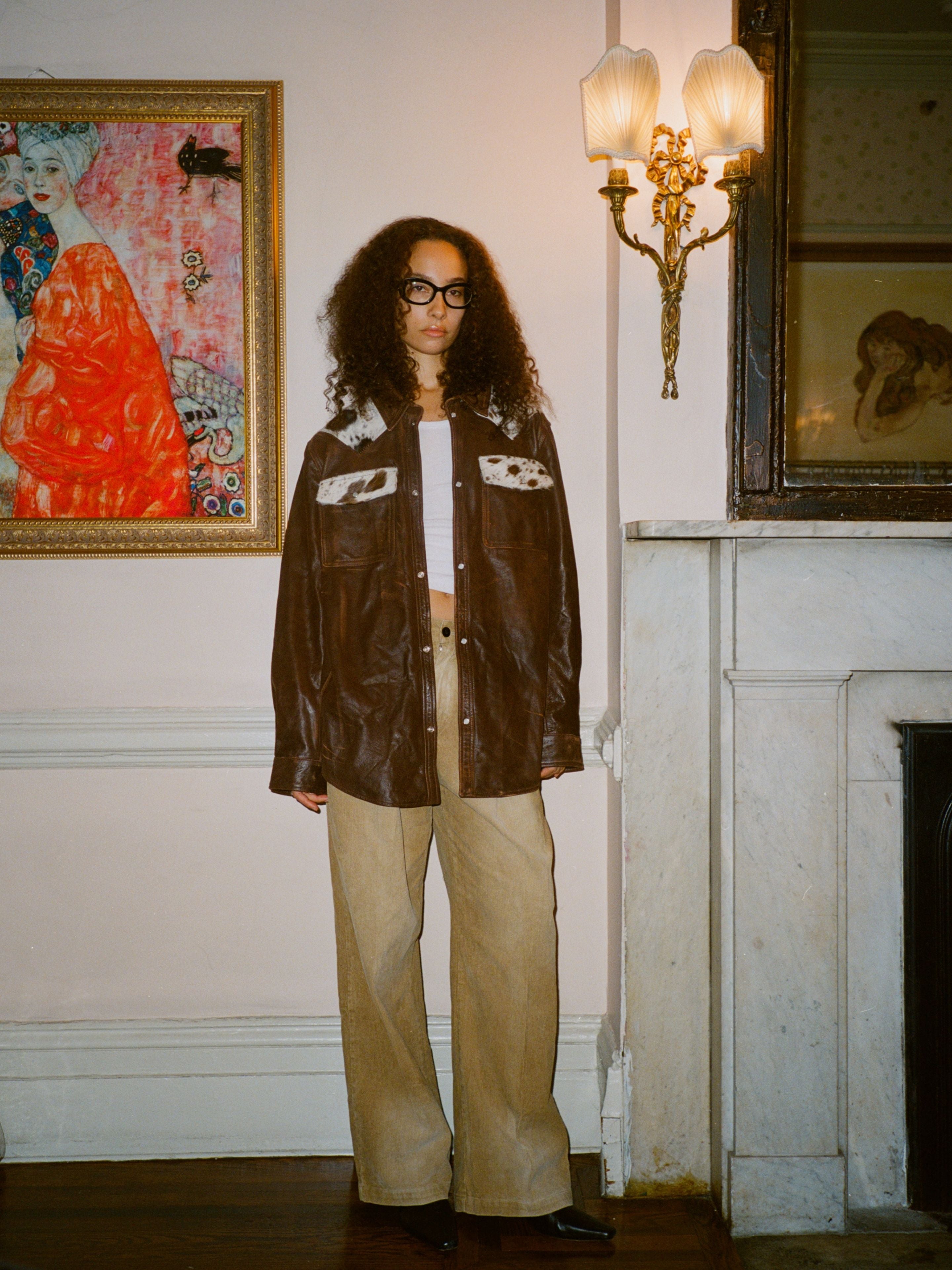 A person with curly hair and glasses stands indoors by a marble fireplace, wearing the FOUND Distressed Leather Cowhide Contrast Western Shirt, beige wide-leg pants, and black shoes.