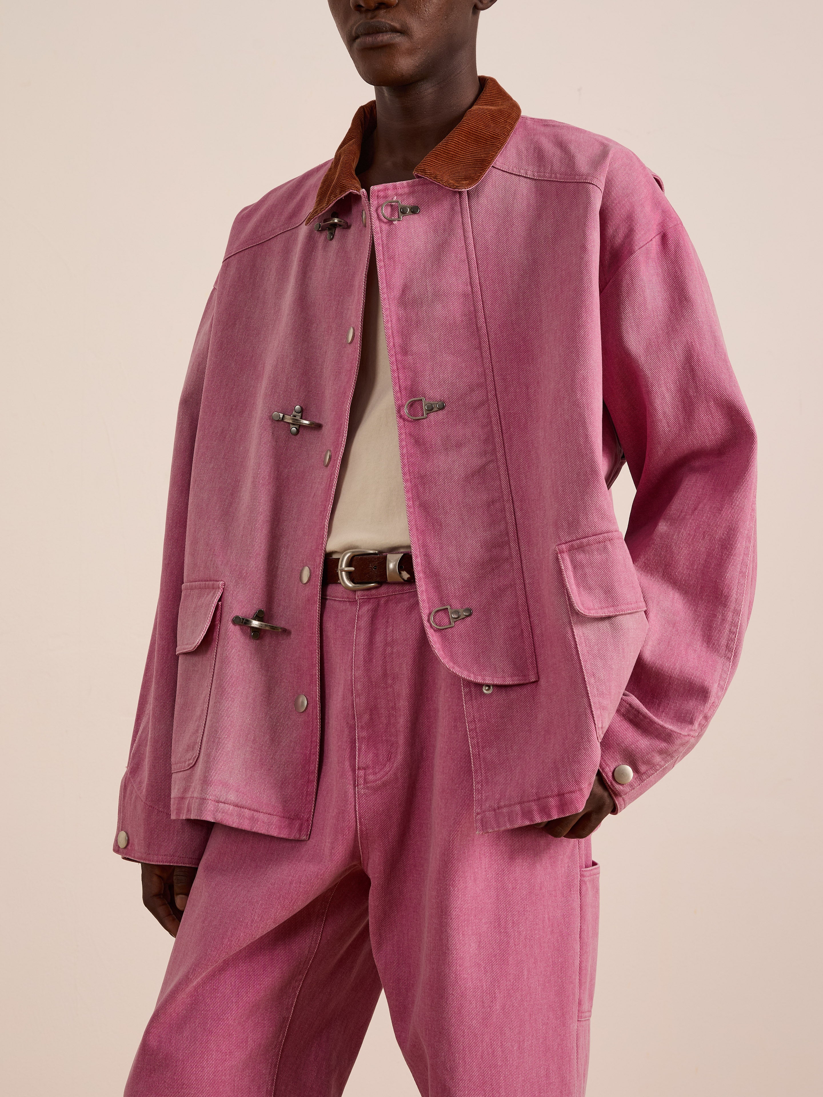 A person wearing Found’s Washed Fireman Clasp Workshop Jacket in pink denim with a brown corduroy collar, styled with a white shirt, pink denim pants, and a brown belt, stands against a plain light background.