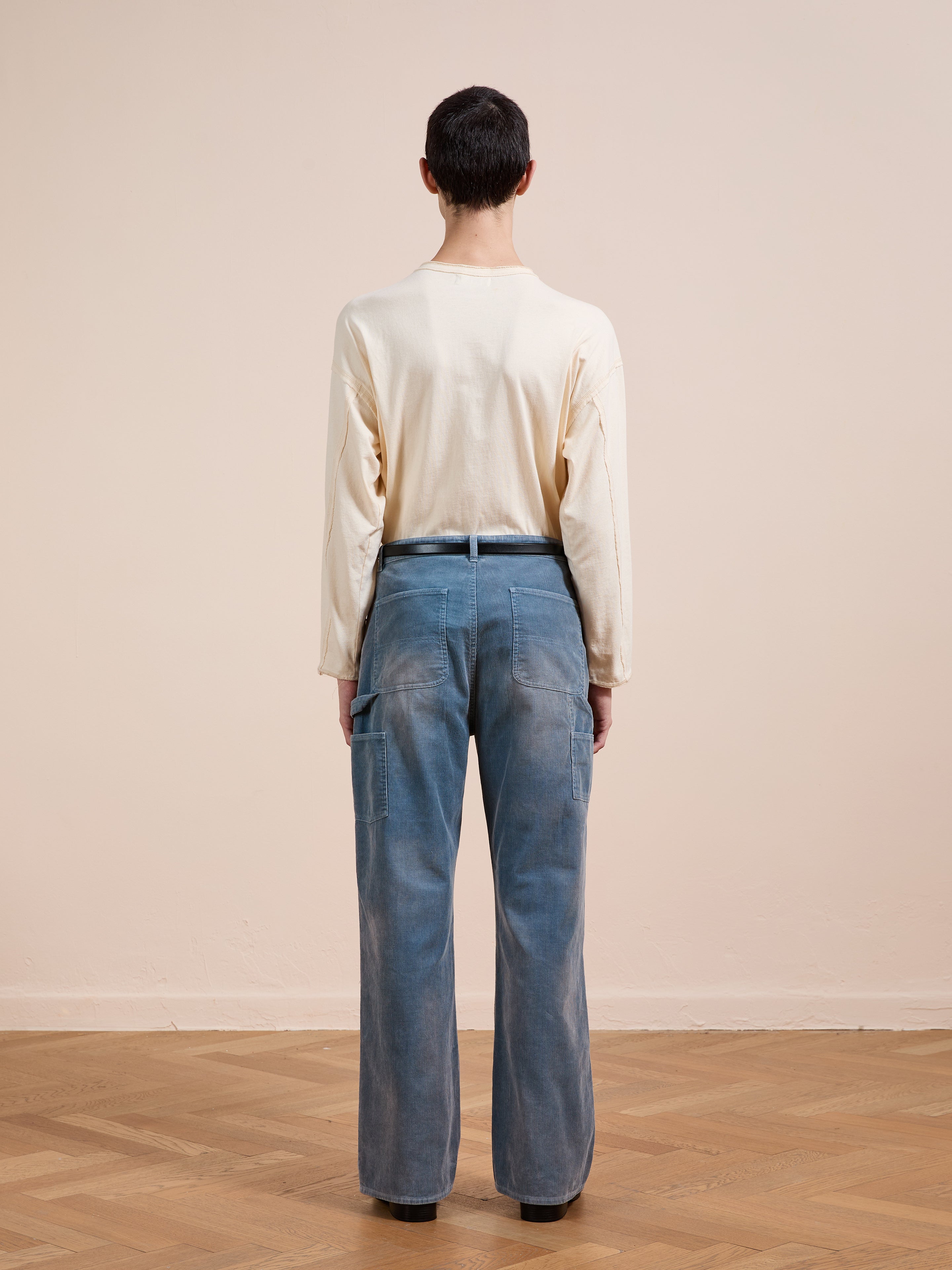 A person stands indoors with their back to the camera, wearing a long-sleeved beige shirt and FOUND Corduroy Flare Work Pants in vintage blue, relaxed fit, on a wooden floor against a plain light wall.