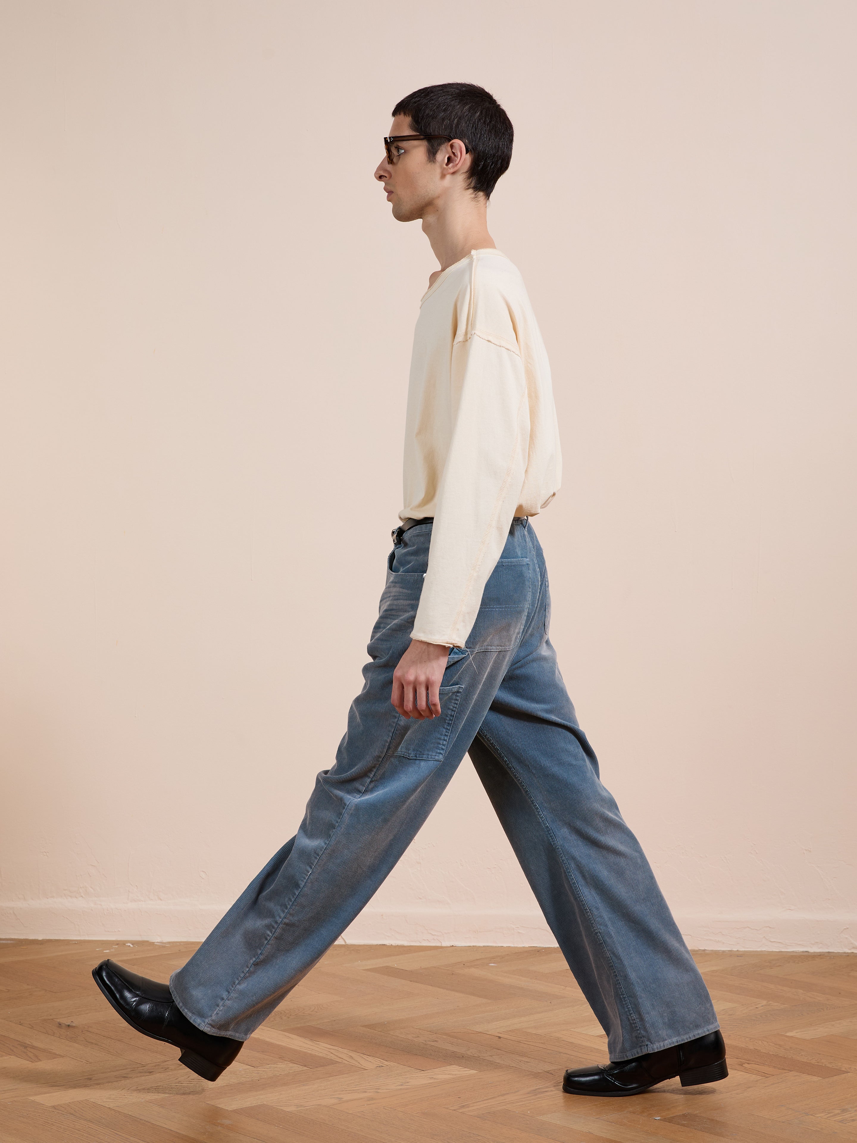 Someone wearing FOUND Corduroy Flare Work Pants in vintage blue, a cream long-sleeve shirt, and black shoes walks indoors on a wooden floor against a plain beige wall.