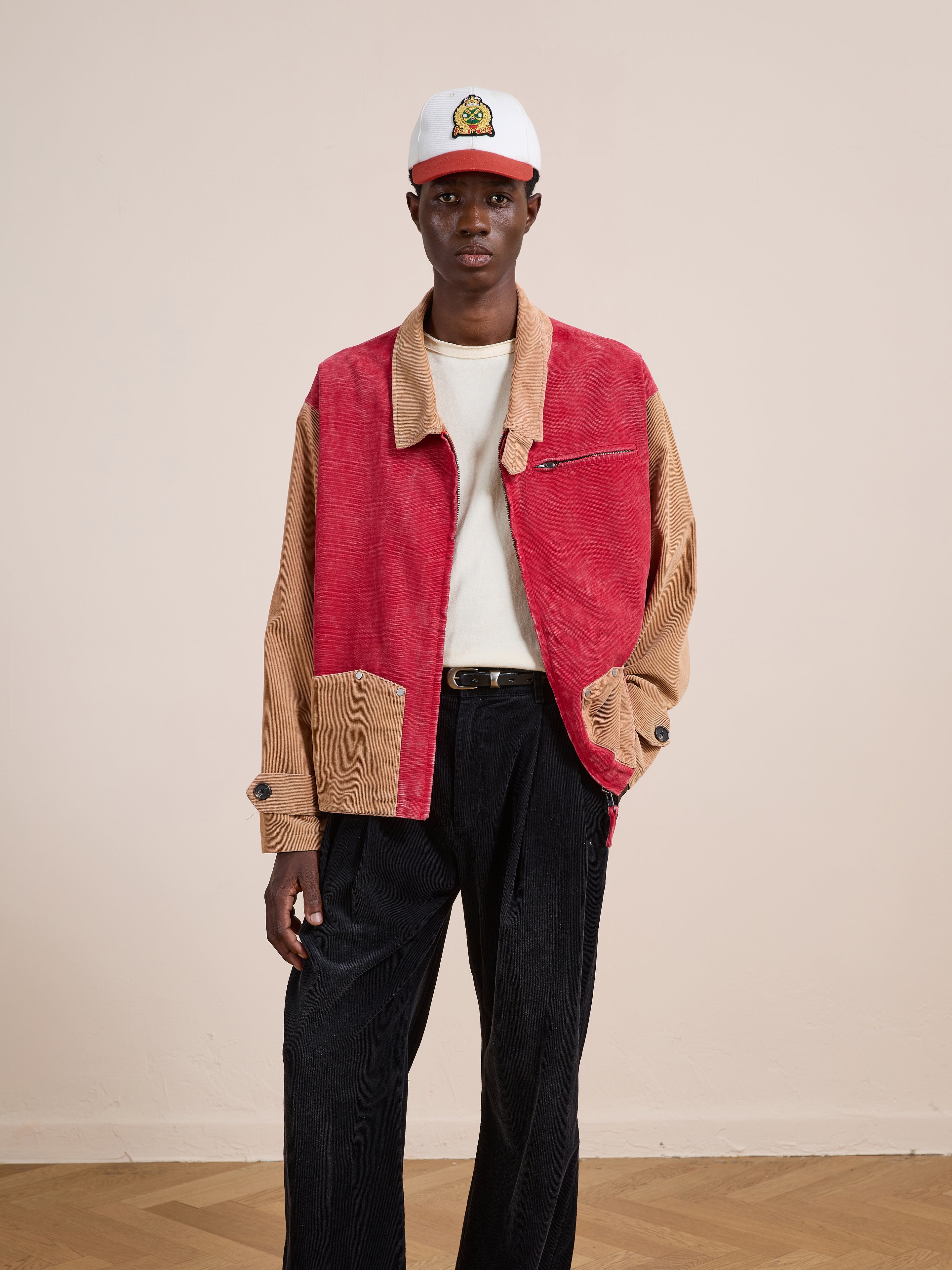 A person stands indoors in a Found Corduroy Field Jacket (unisex, relaxed fit, tan and red), white cap and shirt, and black pants against a plain wall and wooden floor.