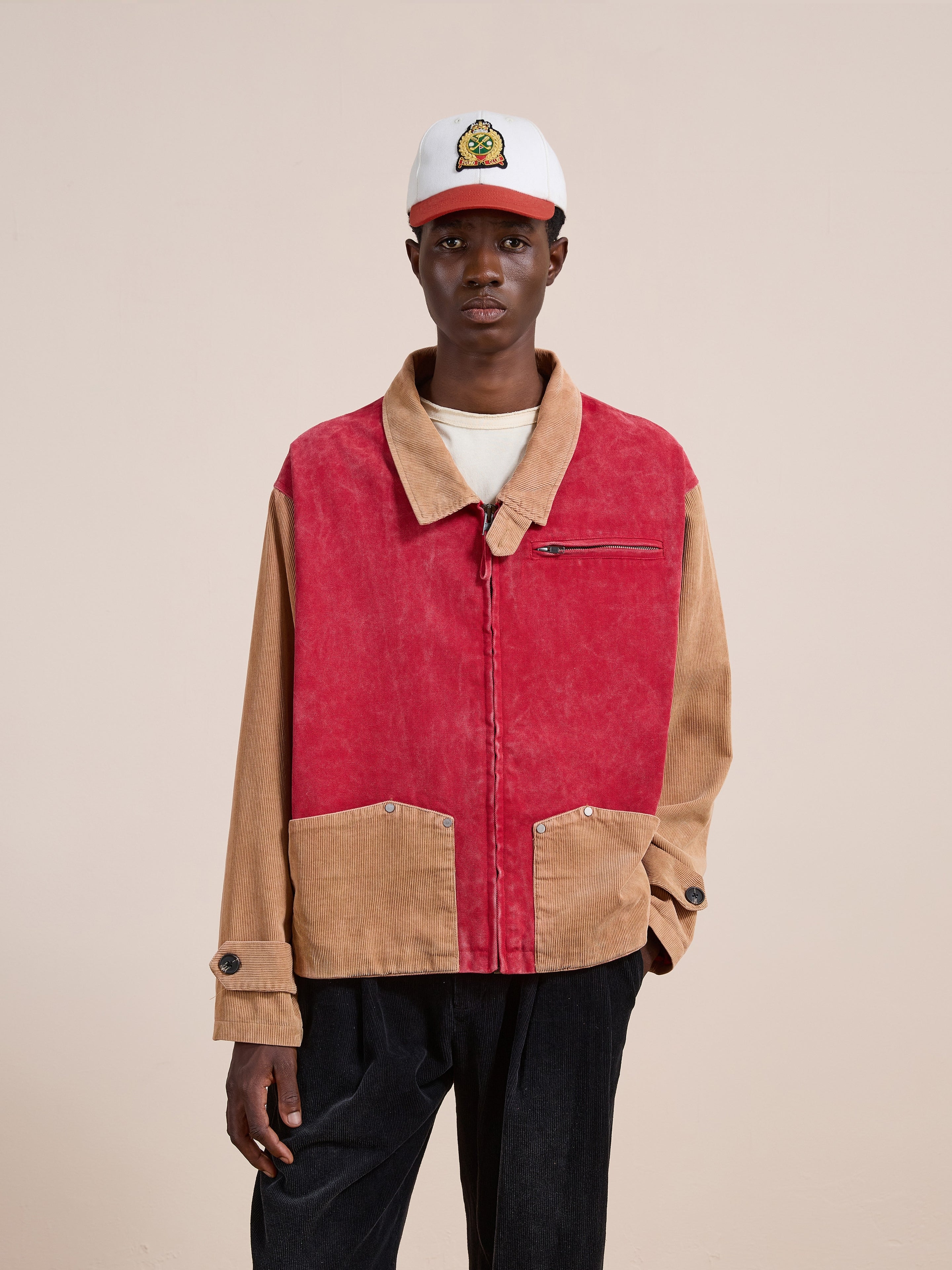 A person wearing the Found Corduroy Field Jacket in red and tan with a relaxed fit, styled with a white T-shirt, black pants, and a white baseball cap featuring a cartoon graphic, stands against a plain beige background.