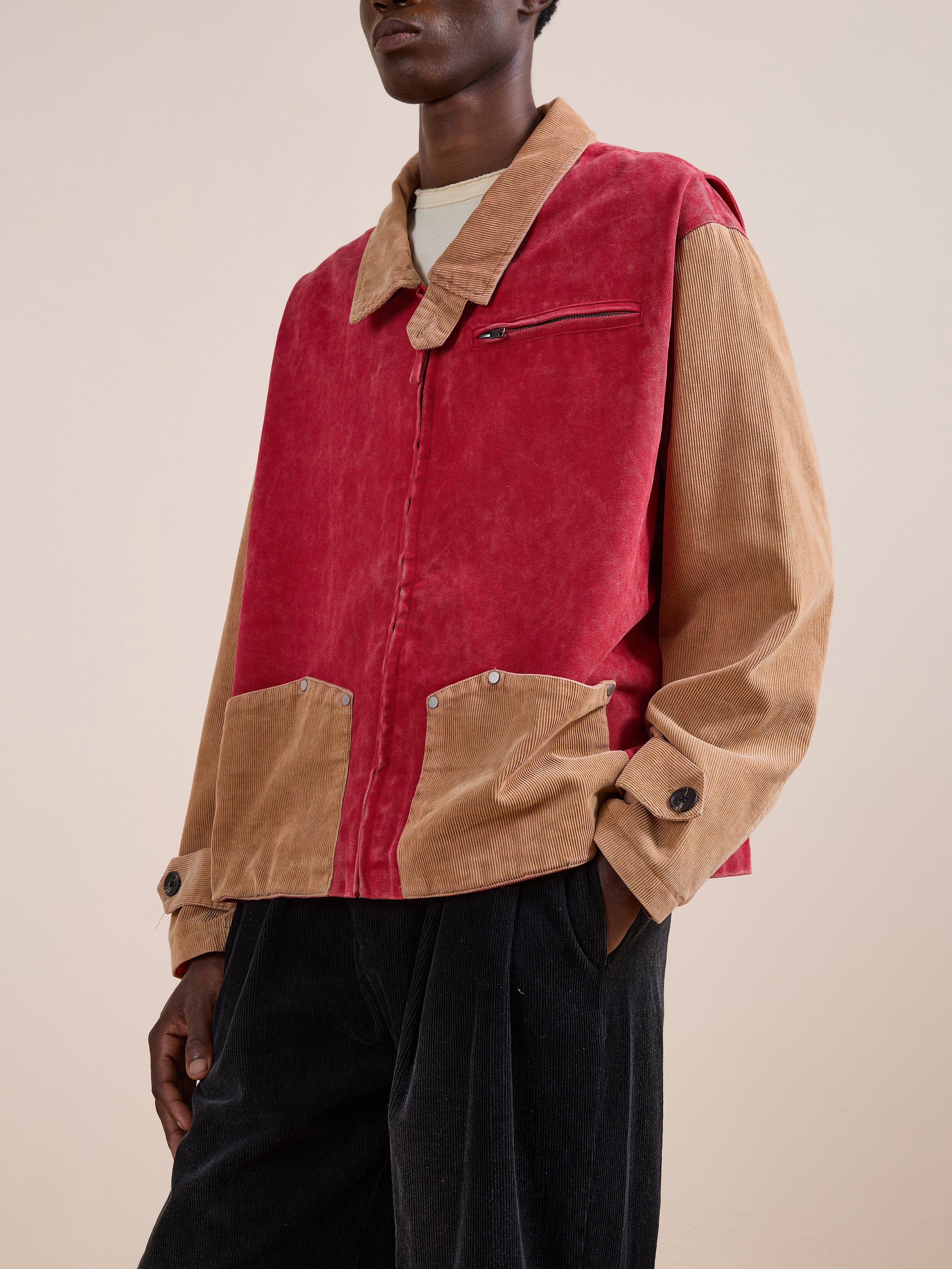 A person wears the Found Corduroy Field Jacket, a color-blocked unisex style with a red front and tan sleeves and pockets, paired with black pants, standing against a plain background.