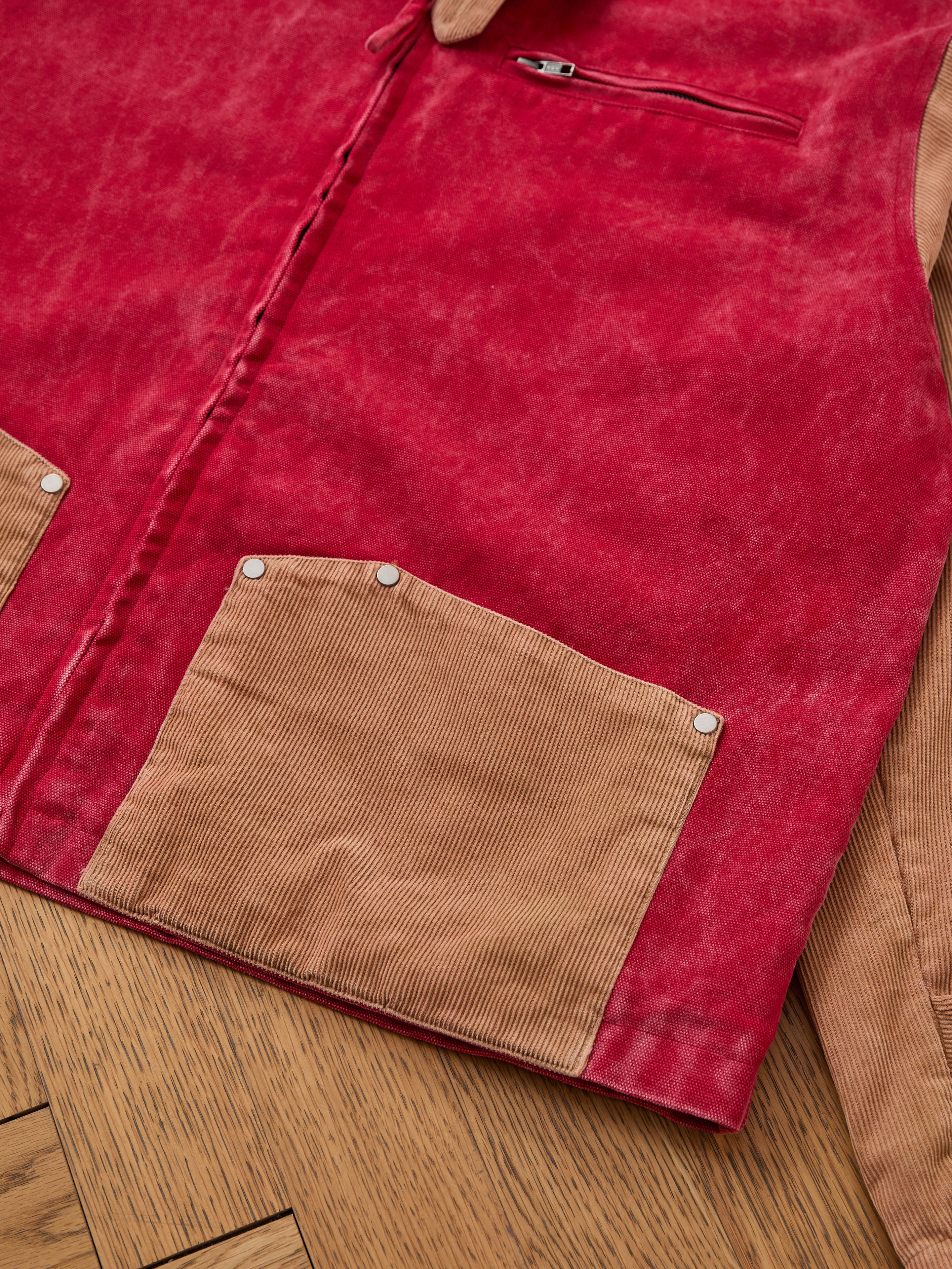 A close-up of Found’s Corduroy Field Jacket in red with tan pockets and sleeves, silver rivets, and a unisex workwear design, laid flat on wood—blending rugged style with practical details.