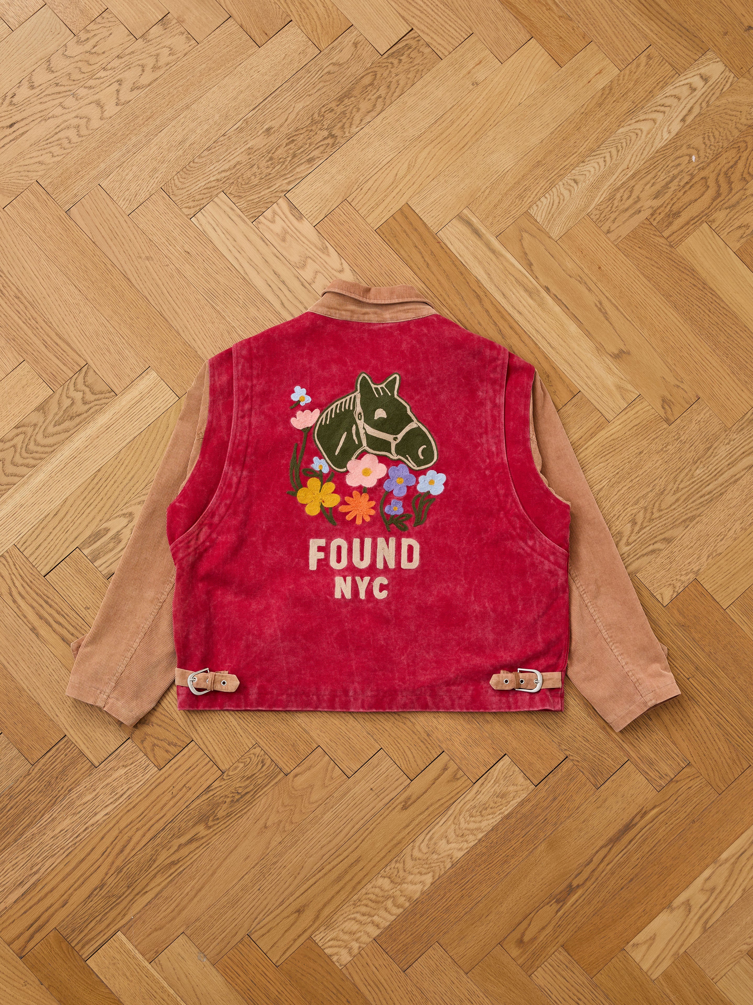 The Found Corduroy Field Jacket, unisex in red and pink, is shown flat on a wooden floor with a horse, flowers, and "FOUND NYC" embroidered on the back—a stylish modern twist on classic workwear.