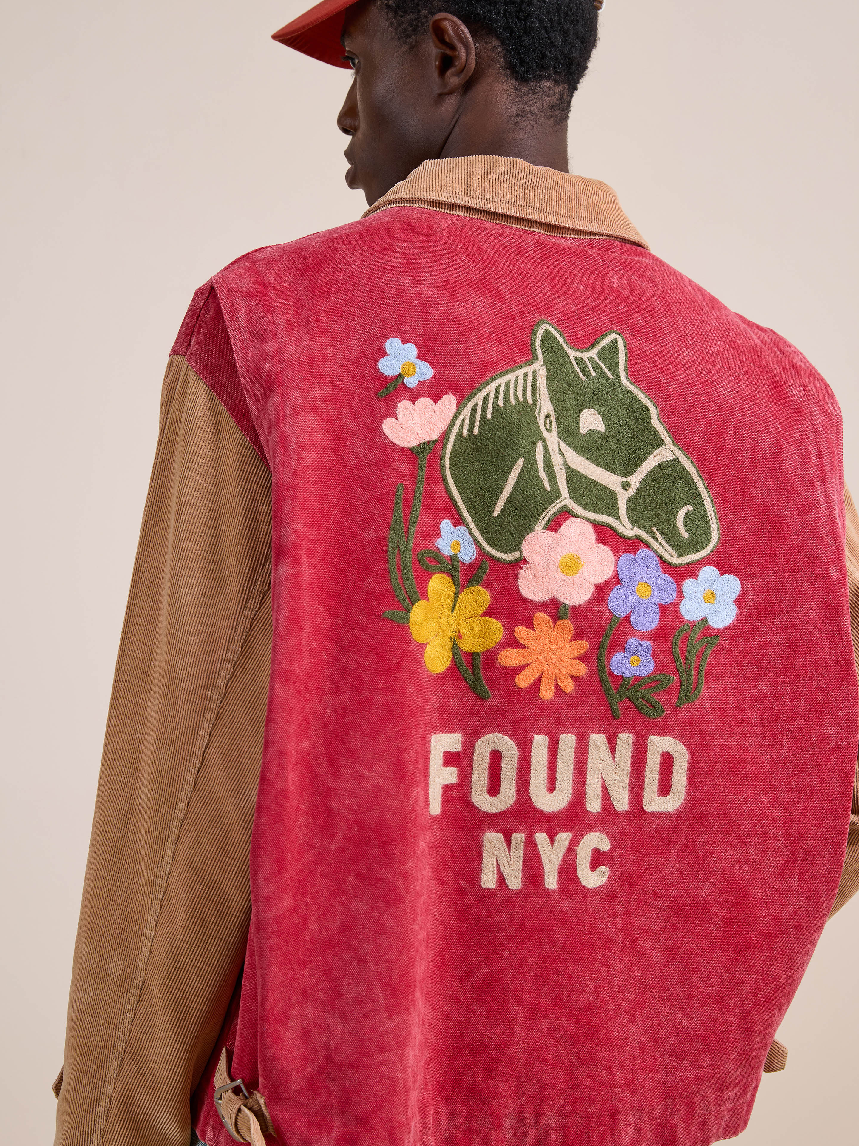 Someone wears the Found Corduroy Field Jacket, a unisex red and tan piece featuring a horse head and colorful flowers embroidered on the back, plus "FOUND NYC" text.