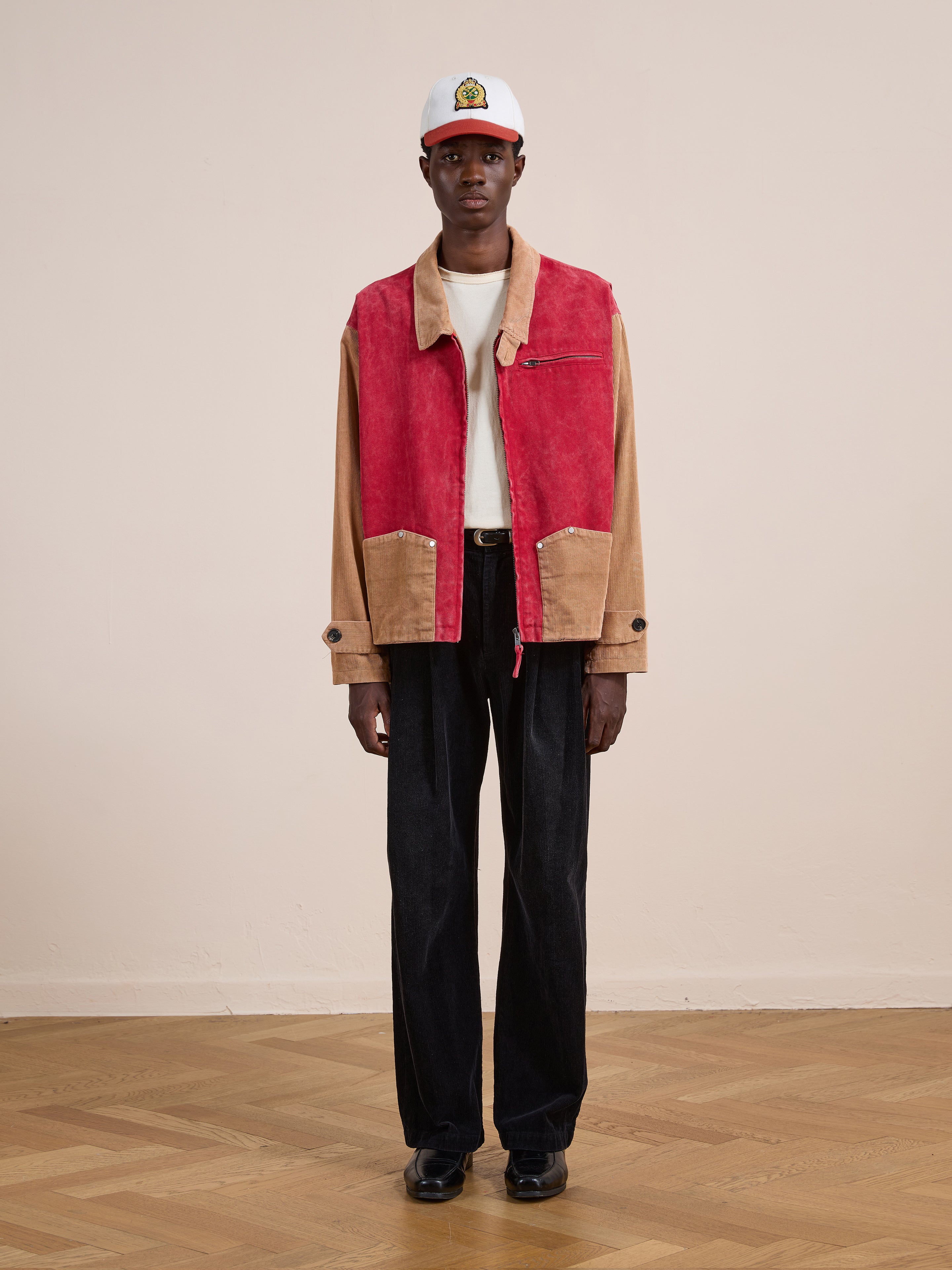 Indoors on a wooden floor, a person wears a white cap, Found’s Corduroy Field Jacket in red and tan panels, a white T-shirt, black trousers, and black shoes.