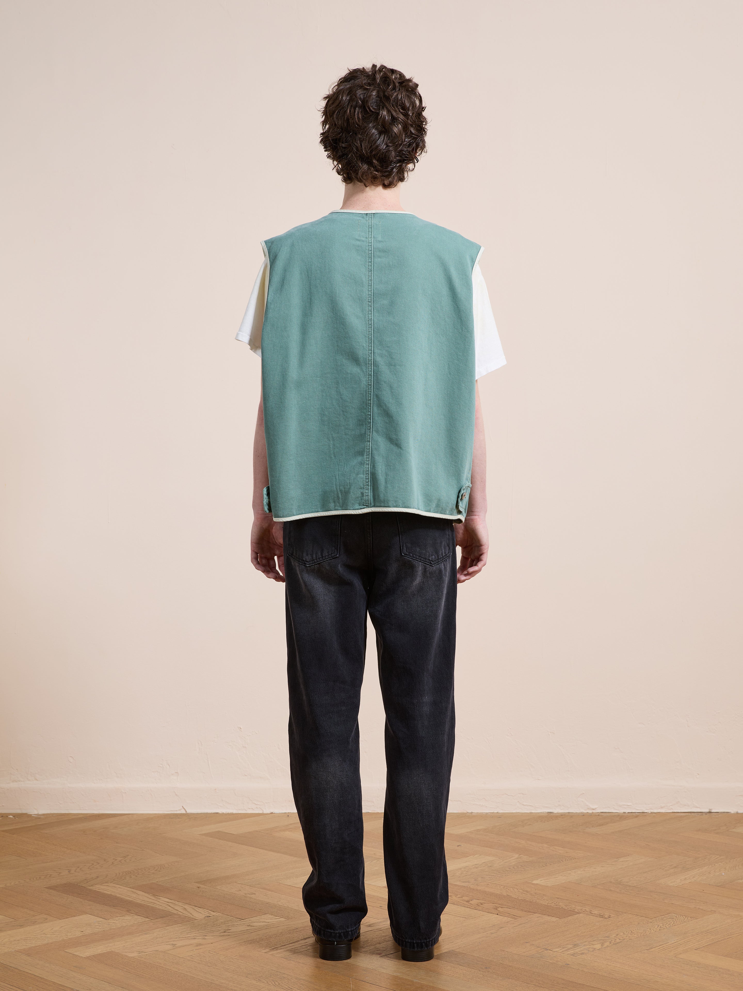 A person stands indoors on a wooden floor, facing away, wearing the FOUND Cotton Twill Utility Vest in vintage washed green over a white shirt and black jeans.