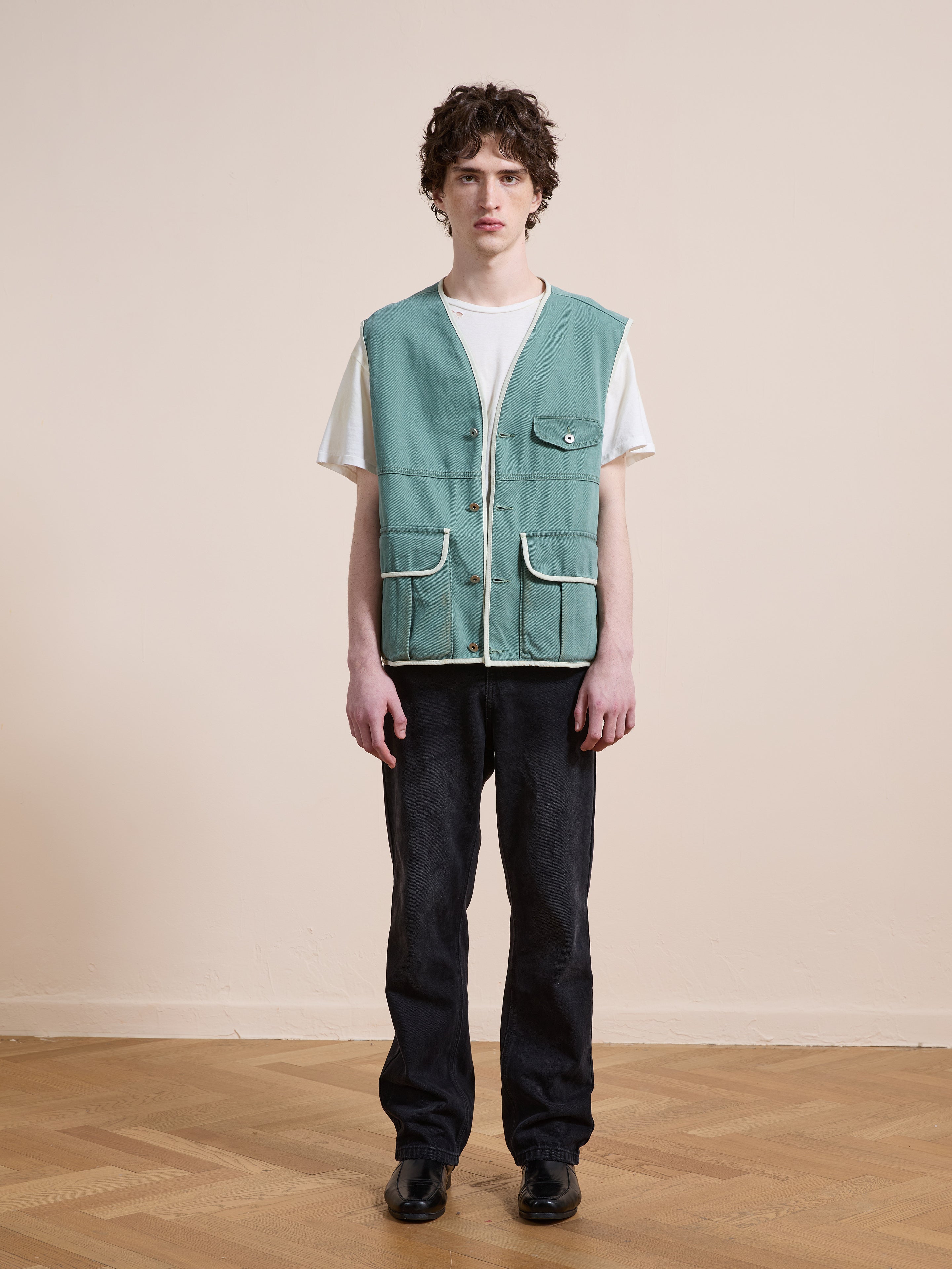 A young person stands indoors wearing the FOUND Cotton Twill Utility Vest in vintage washed green over a white t-shirt, paired with dark jeans and black shoes, arms relaxed at their sides.