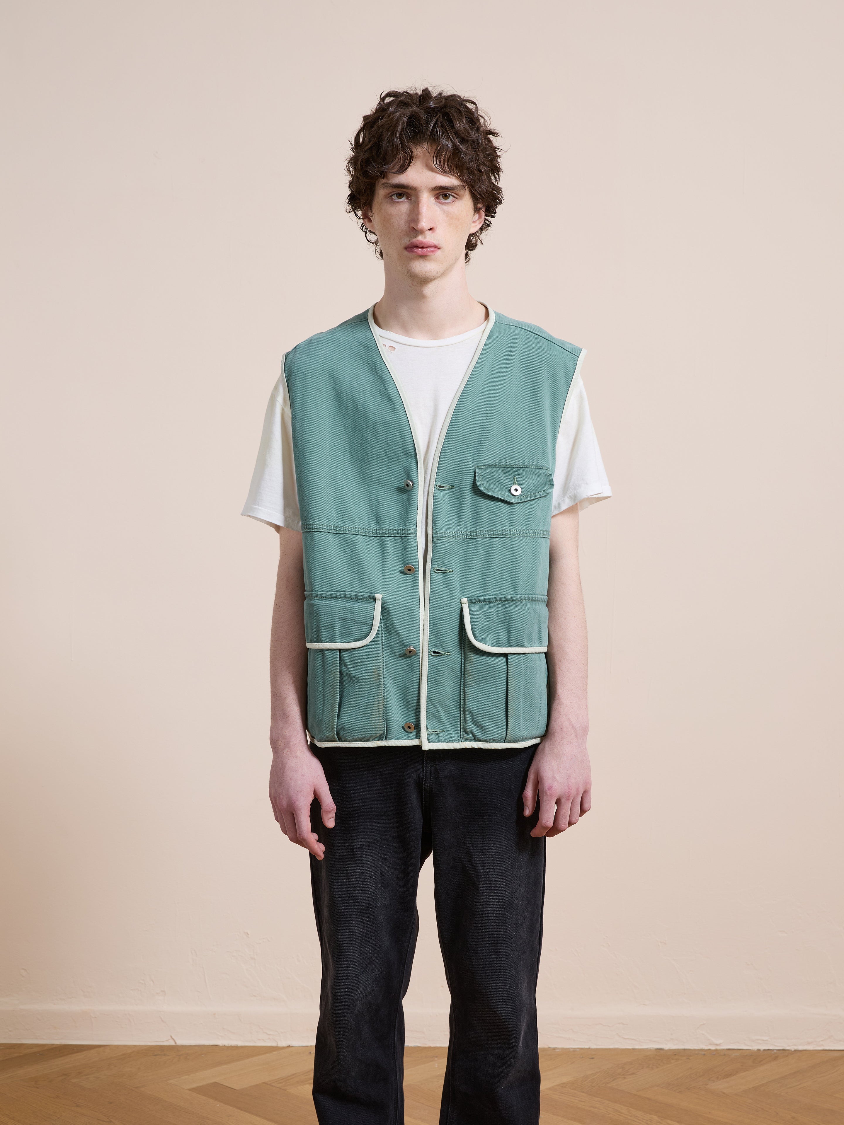 A person stands indoors against a beige wall, wearing a white t-shirt, black pants, and the FOUND Cotton Twill Utility Vest in vintage green with white trim and pockets.