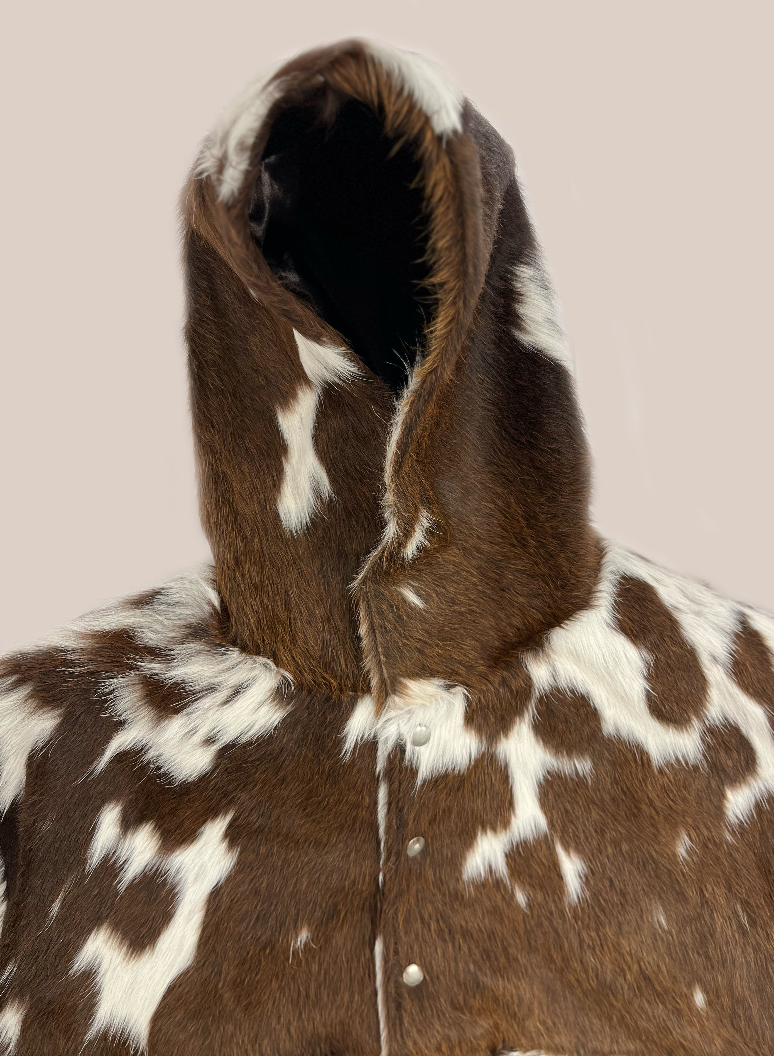 The FOUND Cowhide Drover Hoodie is a unique unisex coat featuring brown and white cowhide-patterned fur, snap buttons, and a hood—shown against a plain background.