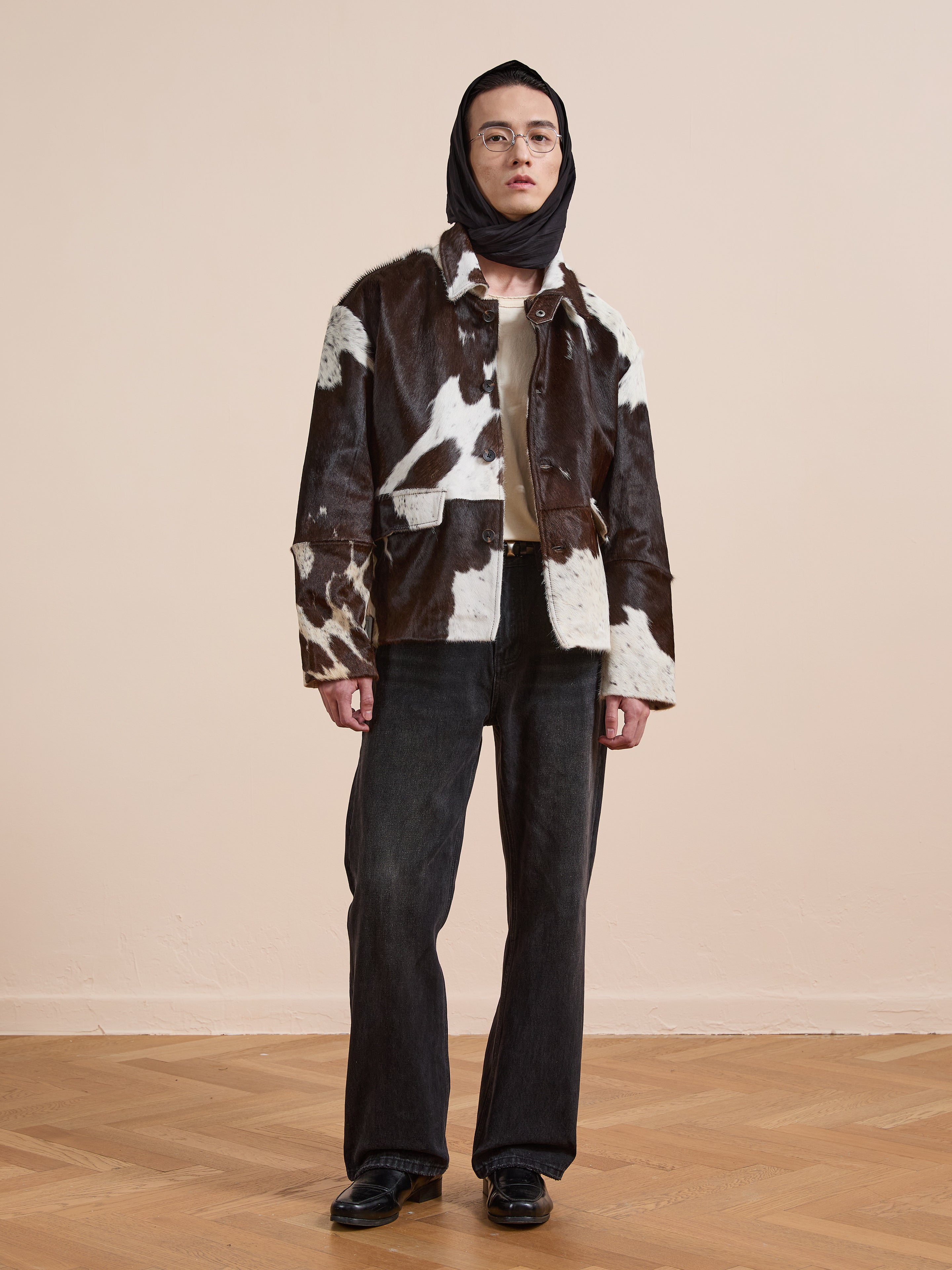 Indoors, a person wears the FOUND Cowhide Prairie Utility Jacket in black and white, styled with black jeans, black shoes, glasses, and a black scarf covering their head.