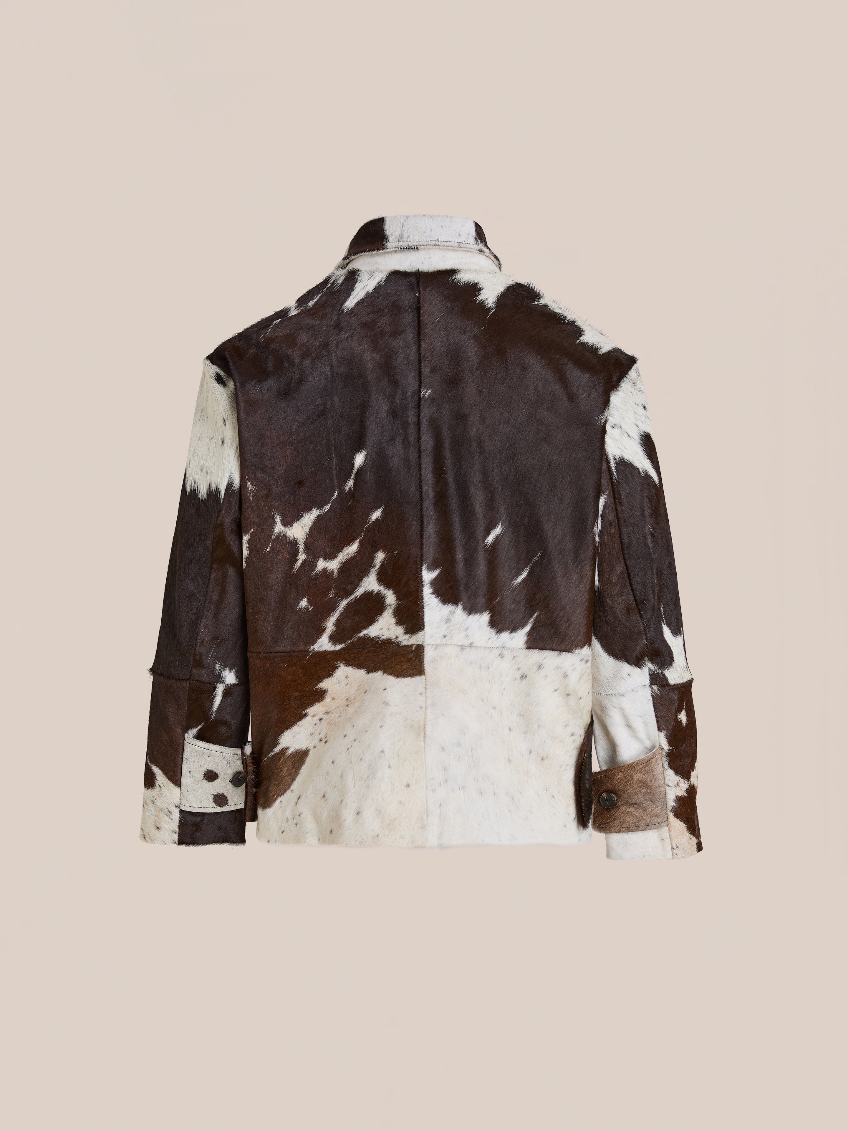 A rear view of the FOUND Cowhide Prairie Utility Jacket, crafted from genuine leather with a distinctive brown and white spotted pattern, displayed against a plain beige background.
