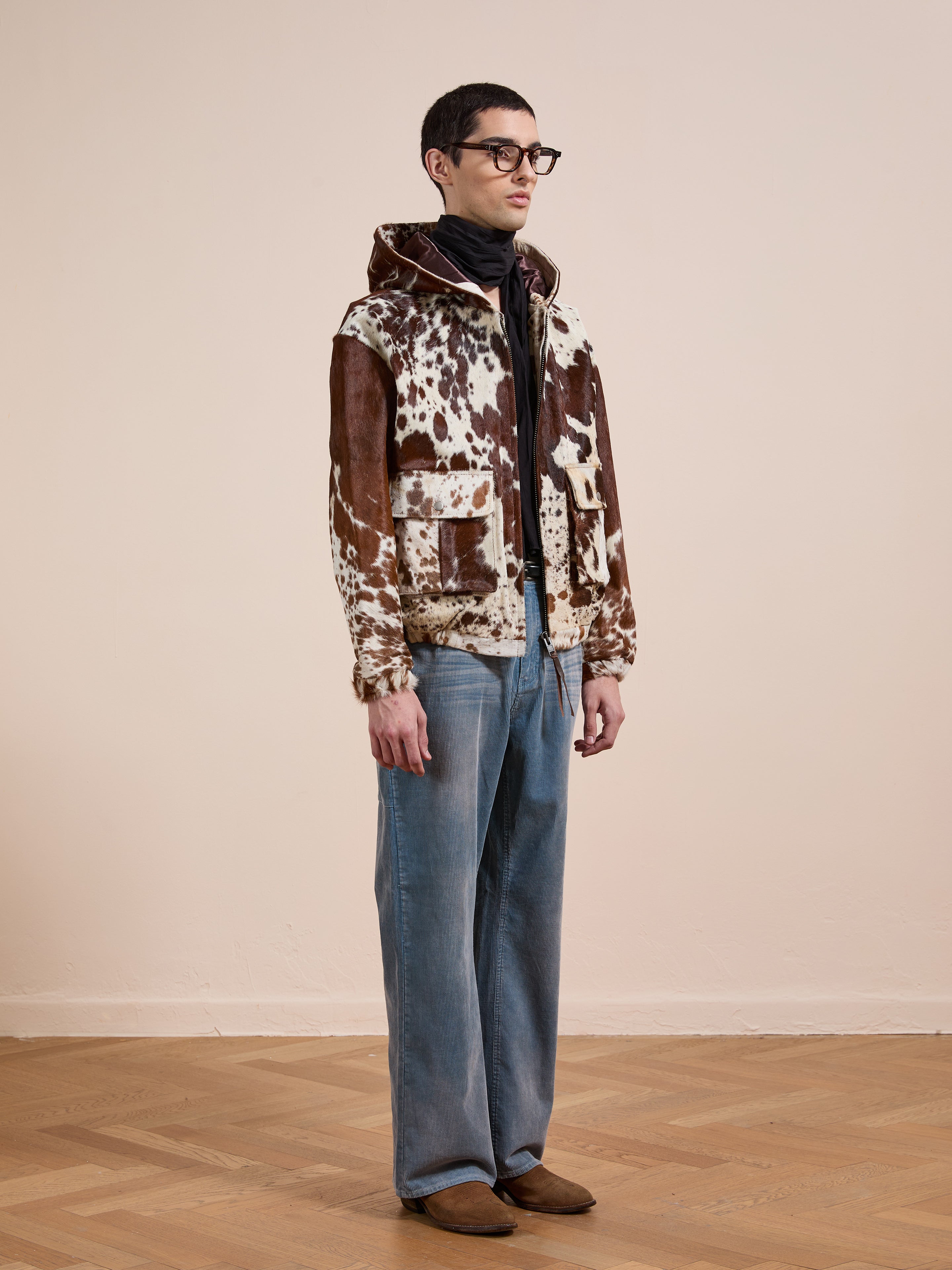 Indoors, a person wears FOUND’s Cowhide Drover Hoodie with brown and white patterns, paired with glasses, a black scarf, blue jeans, and brown shoes on a wooden floor.