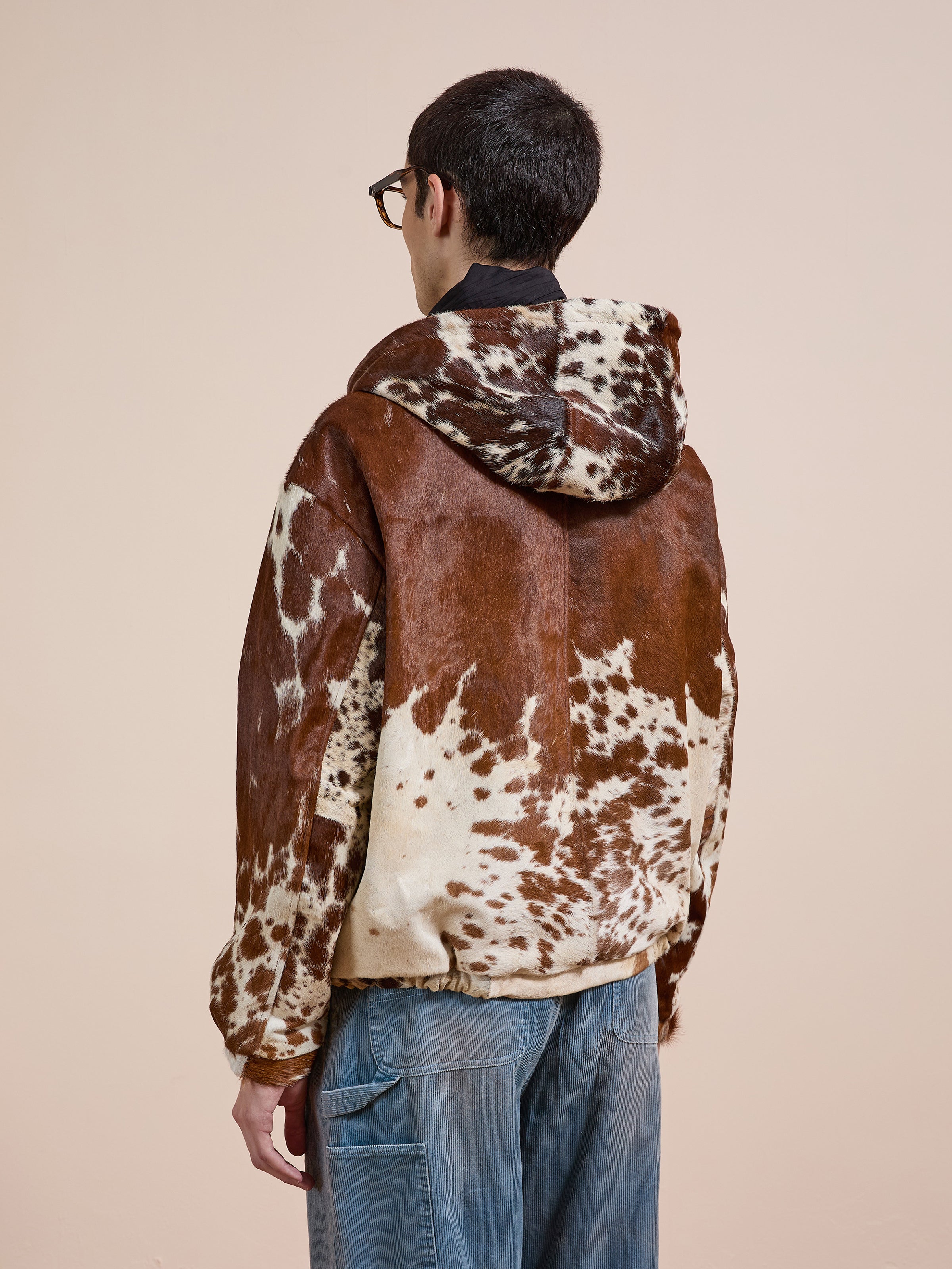 Someone models the FOUND Cowhide Drover Hoodie, featuring a brown and white cowhide pattern, paired with blue jeans and shown from the back against a plain beige background.