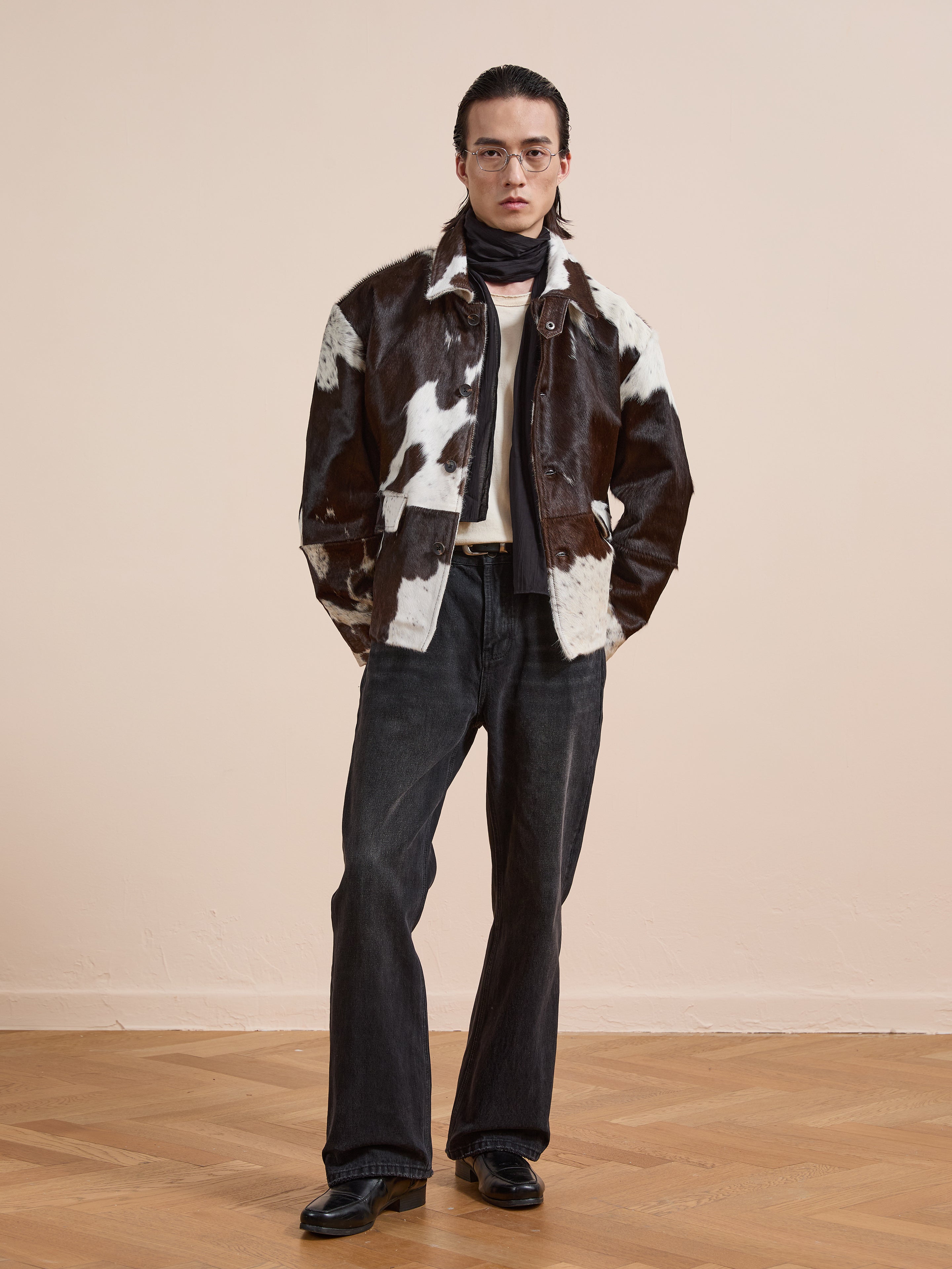 A person wearing glasses, the FOUND Cowhide Prairie Utility Jacket, dark flared jeans, black shoes, and a dark scarf stands indoors on a wooden floor against a plain light wall.