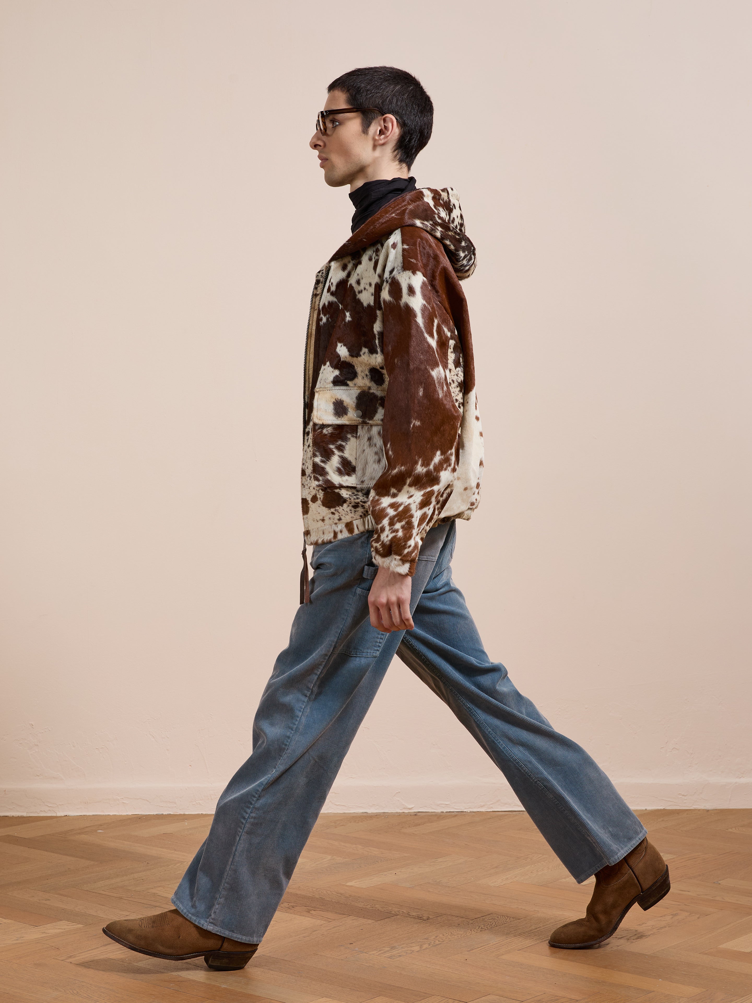 Someone walks indoors wearing the FOUND Cowhide Drover Hoodie, wide-leg blue jeans, brown boots, and glasses against a plain light-colored wall.