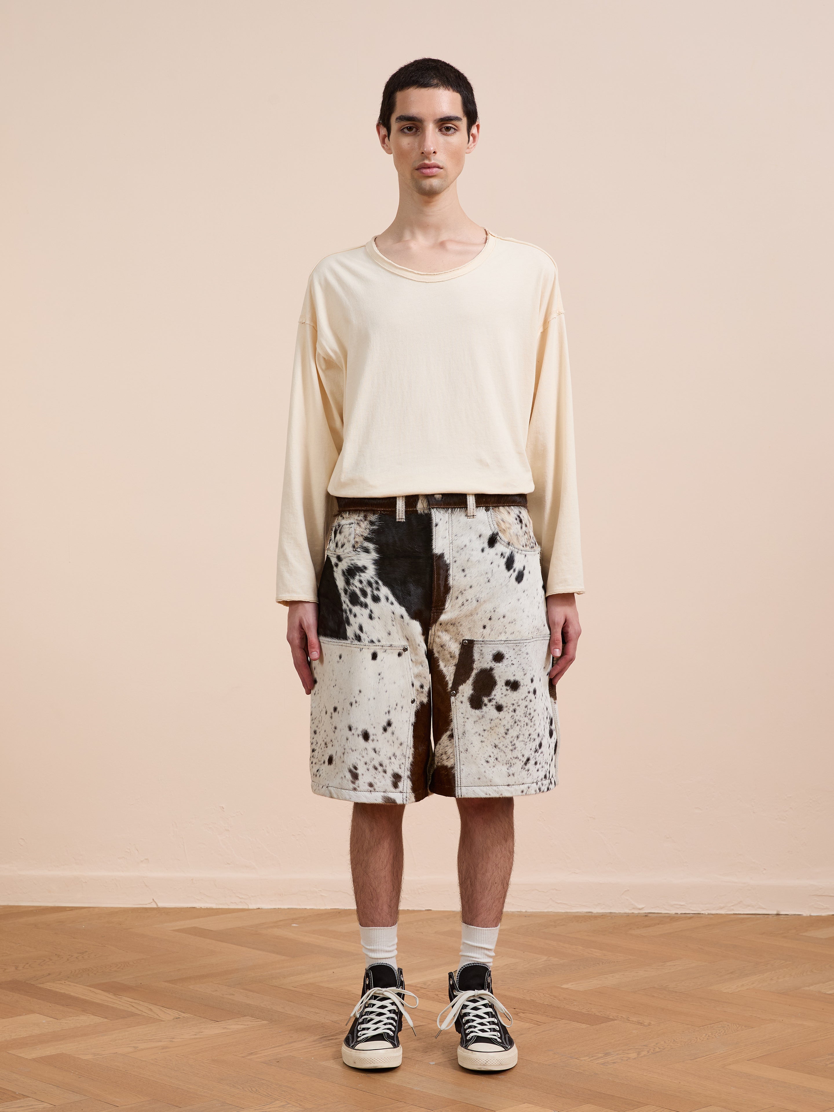 A person stands indoors wearing a loose cream long-sleeve shirt, Found Cowhide Prairie Carpenter Shorts in a black-and-white print, white socks, and black sneakers for a relaxed unisex fit.