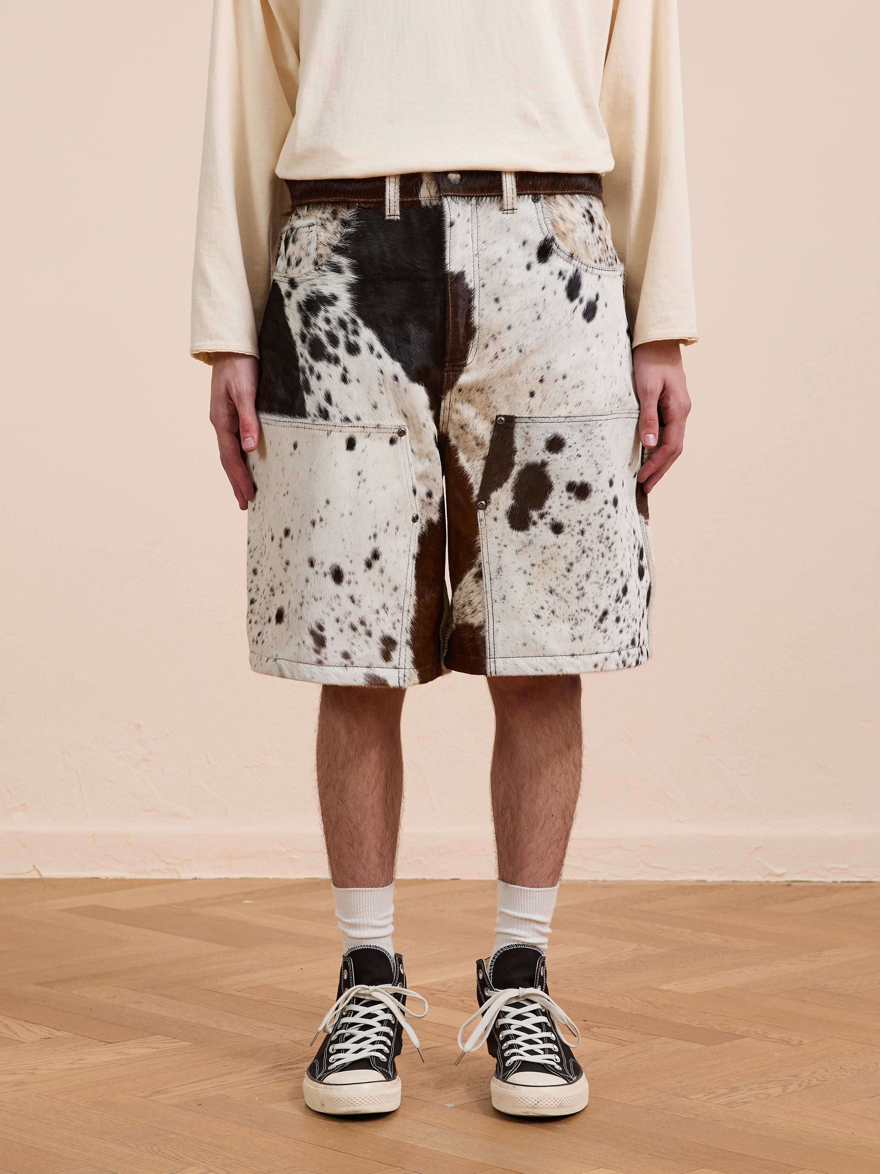A person wearing a long-sleeve cream shirt, Found Cowhide Prairie Carpenter Shorts with large front pockets, white socks, and black sneakers stands on a wooden floor against a beige wall.