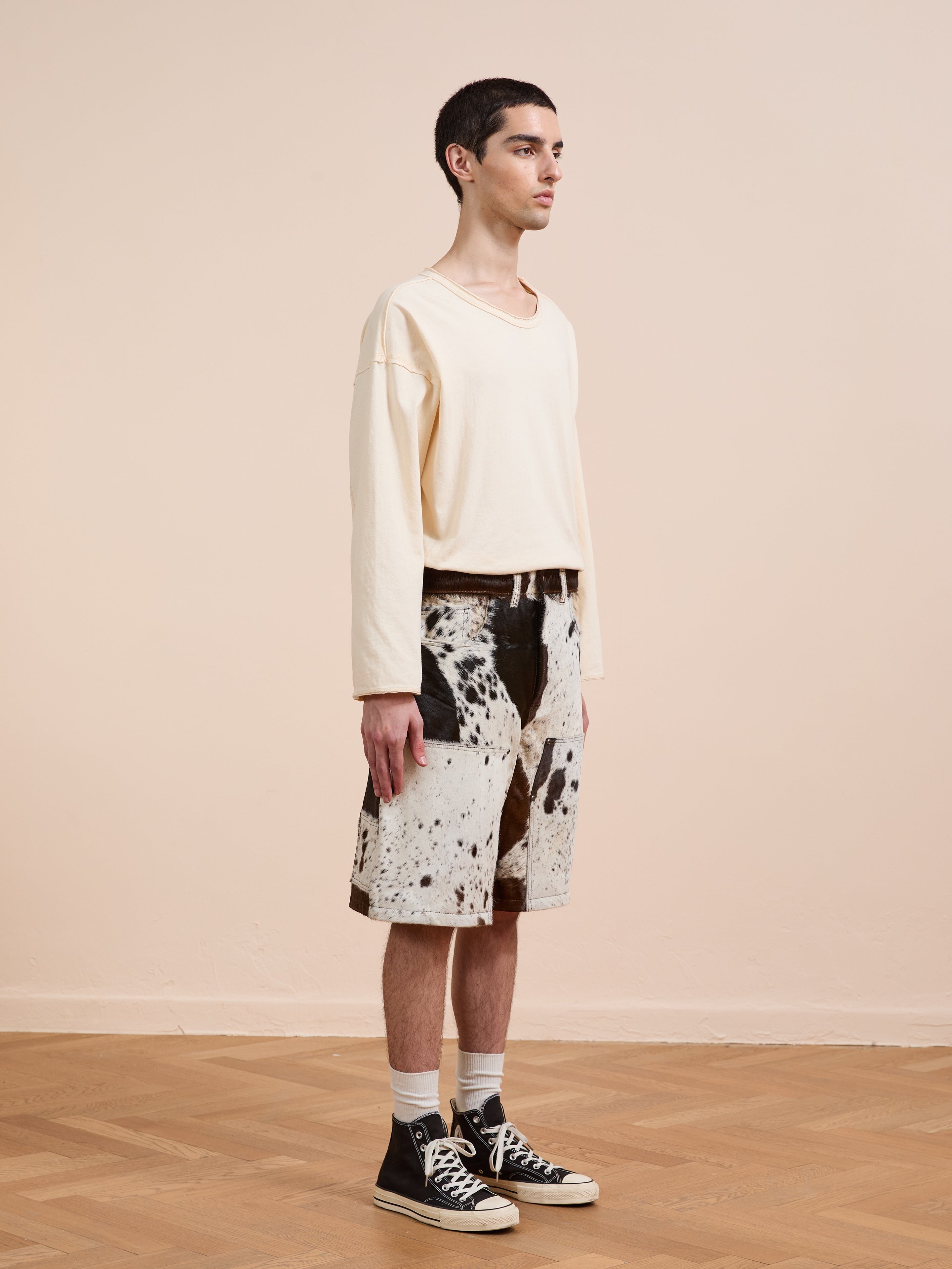 A person stands indoors on a wooden floor, wearing a cream long-sleeve shirt, Found Cowhide Prairie Carpenter Shorts (unisex, relaxed fit), white socks, and black high-top sneakers, facing to the right.