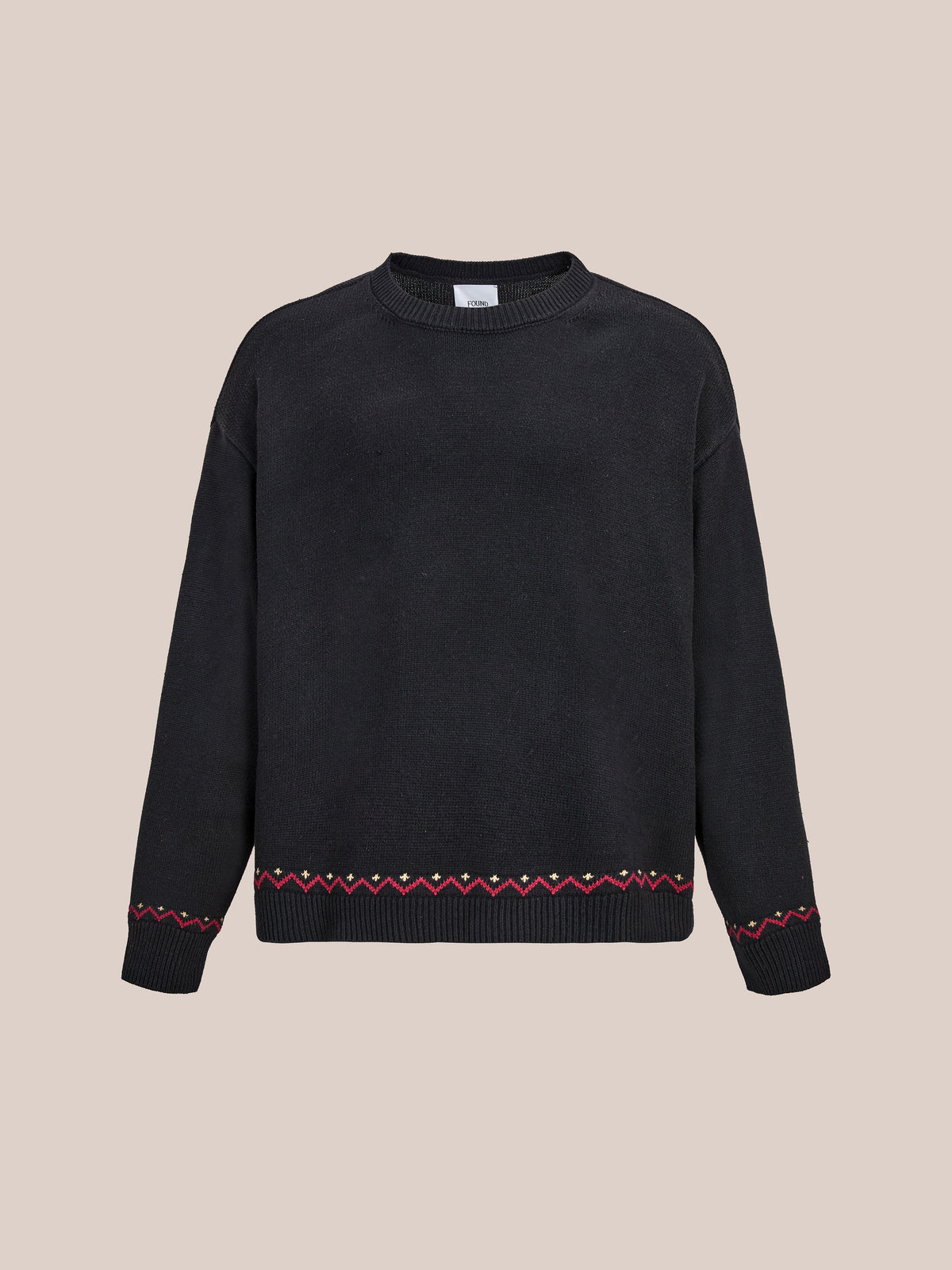 The FOUND Crest Sweater is a black long-sleeve knit with a ribbed collar, cuffs, and red-yellow zigzag trim, shown on a beige background.