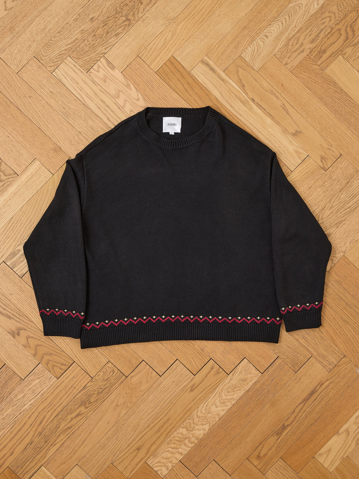 The FOUND Crest Sweater, a black varsity crewneck with red and white zigzag trim on the cuffs and hem, is laid flat on a wooden herringbone floor.