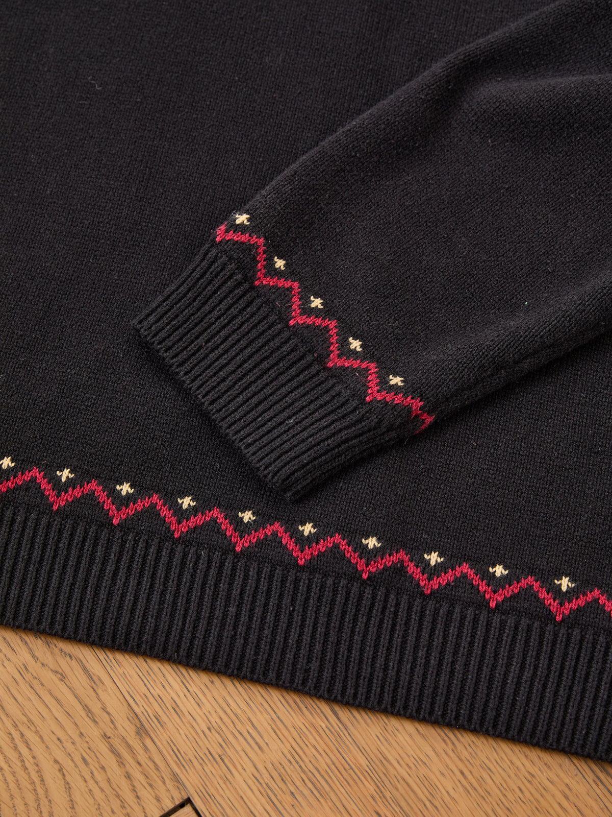 The FOUND Crest Sweater, a black varsity crewneck with ribbed cuffs and hem, features red zigzag and gold dot patterns and is laid flat on a wooden surface.