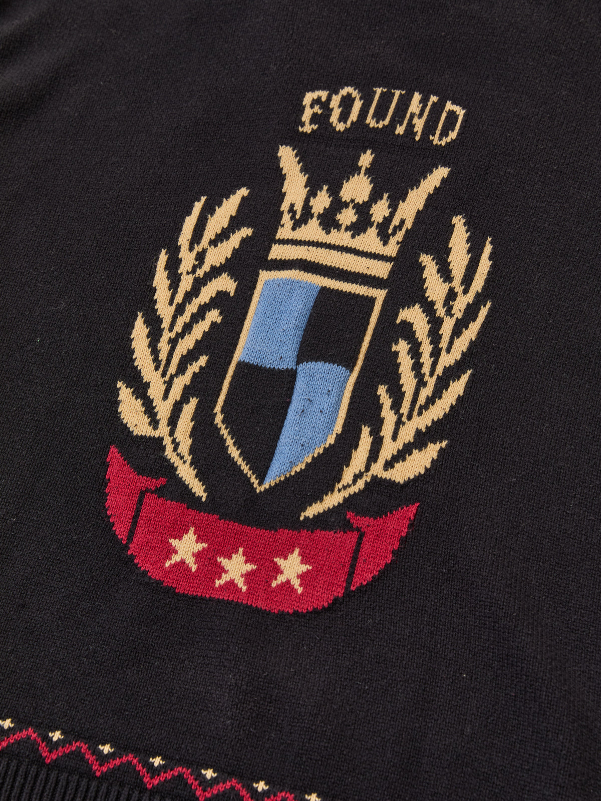The Crest Sweater by FOUND is a black knitwear piece with a stitched crest, crown, laurel branches, shield, “FOUND,” and a red banner with three yellow stars.