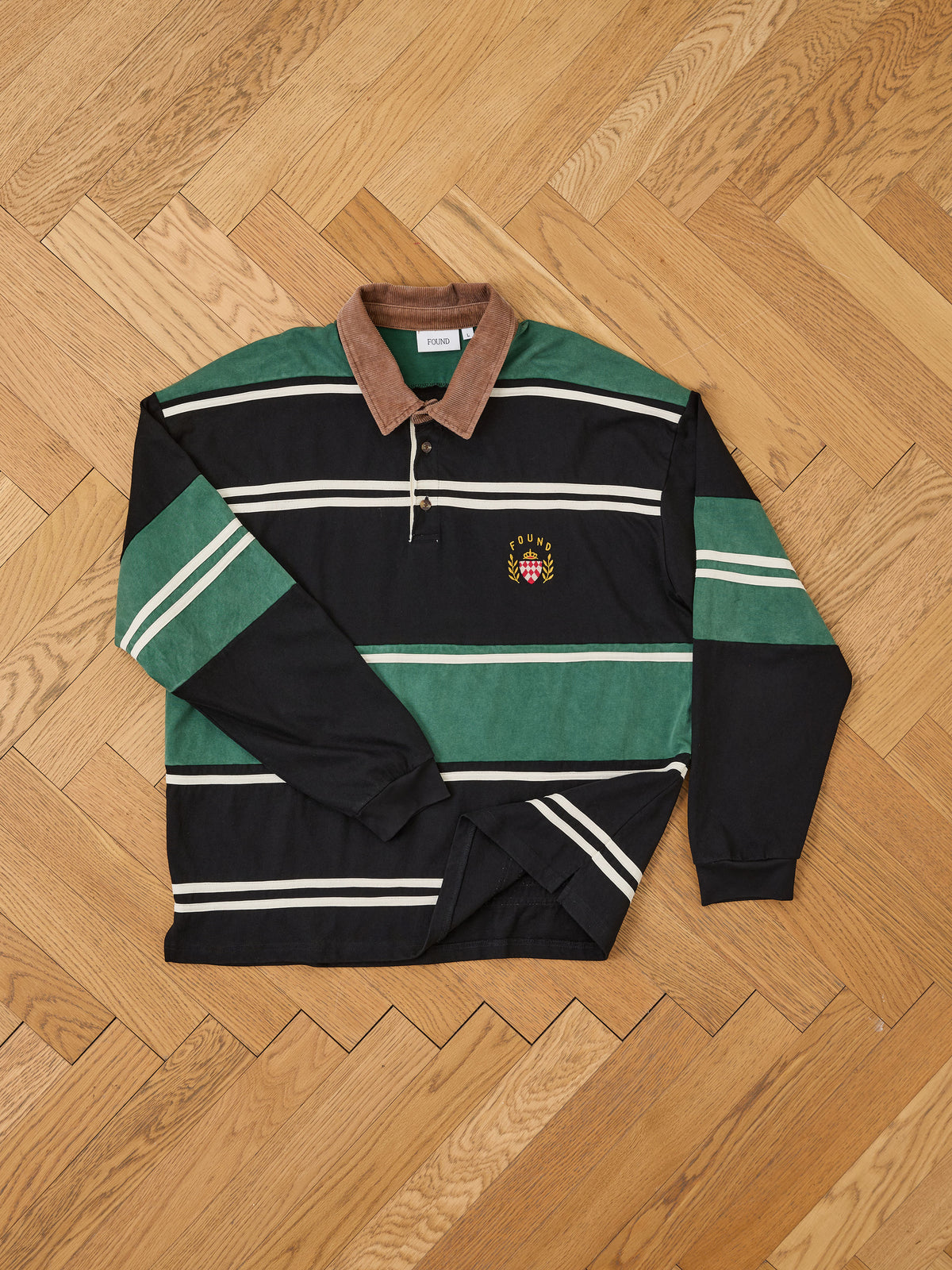 The FOUND Crestfield Rugby is a unisex shirt with green and navy stripes, a brown collar, and an embroidered crest, shown flat on a wooden floor.