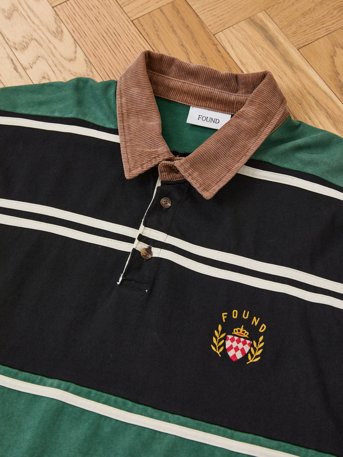 The Crestfield Rugby by FOUND—striped with a brown collar, “FOUND” label, and crest with crown and laurel—is laid flat on a wooden floor.
