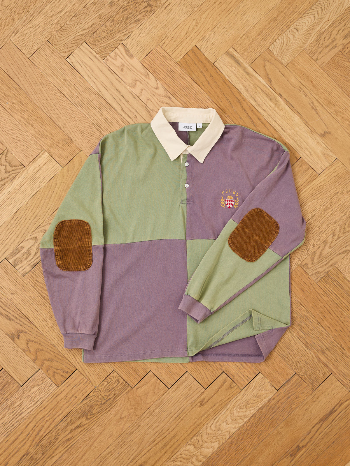 Sure! Here’s your rewritten sentence using the provided product data: The FOUND Crestfield Rugby is a classic athletic rugby-inspired piece, featuring color-blocked green and purple panels, a tan collar, brown suede elbow patches, and an embroidered crest logo. Designed with a unisex relaxed fit, it is pictured laid flat on a wooden parquet floor.