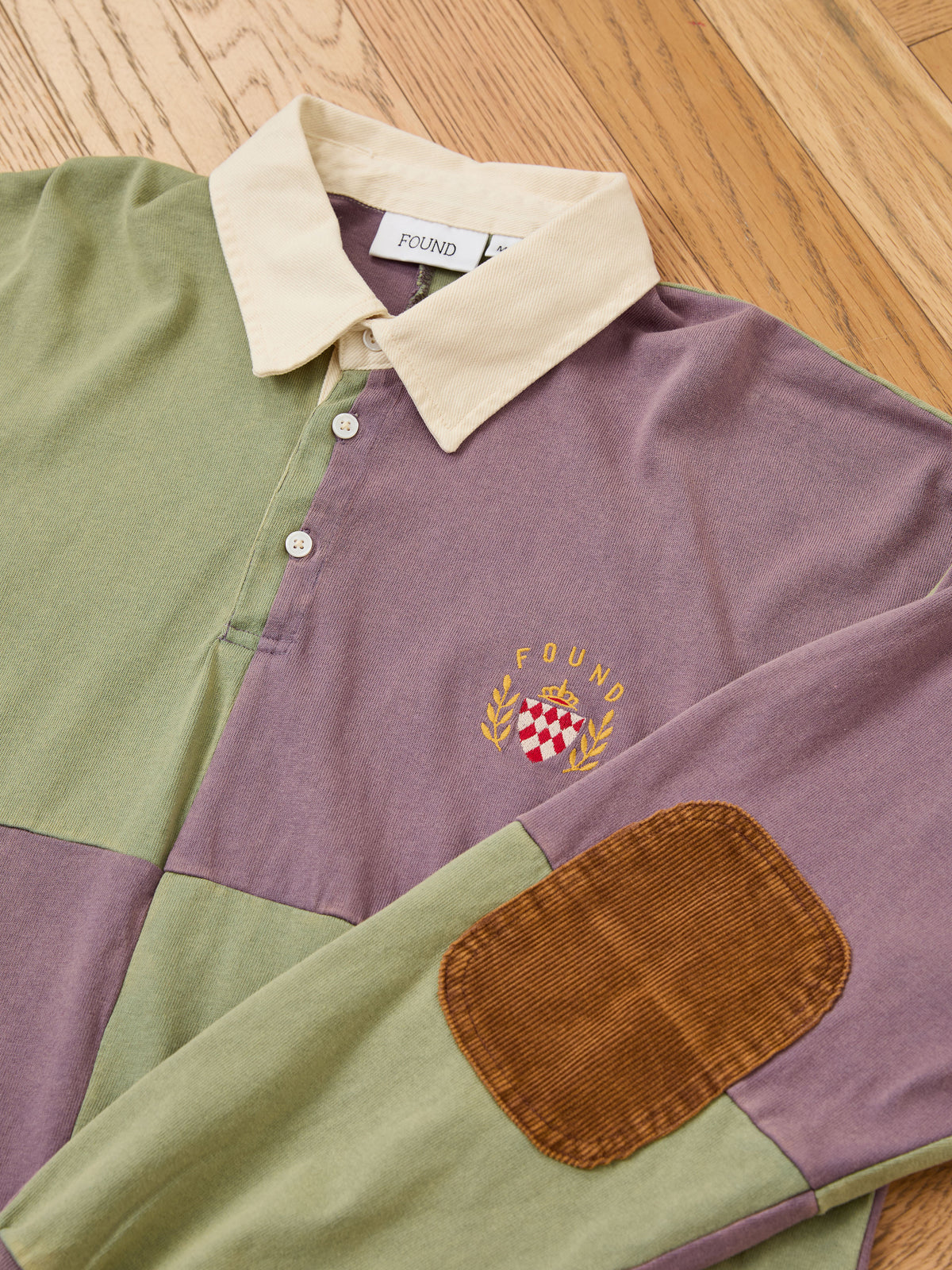 The FOUND Crestfield Rugby, a unisex vintage rugby shirt with a cream collar, embroidered crest, and brown corduroy elbow patches, rests on a wooden floor—seamlessly combining classic style and timeless comfort.