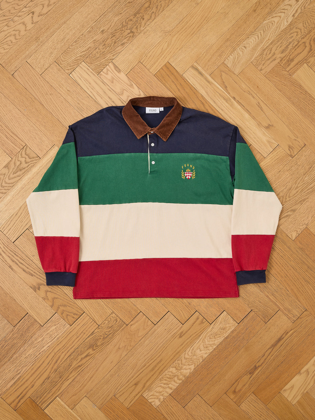 Laid flat on a wood floor, the FOUND Crestfield Rugby showcases long sleeves and bold wide horizontal stripes in navy, green, cream, and red. A brown corduroy collar and an embroidered crest on the green stripe complete this classic unisex rugby style.