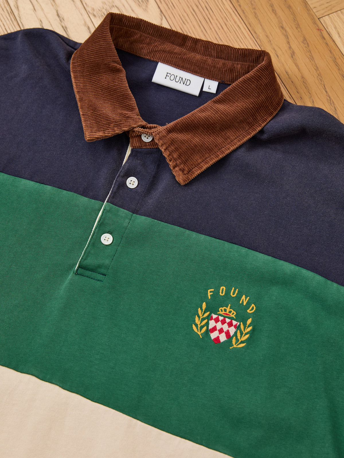 The FOUND Crestfield Rugby is laid flat on a wooden floor, showcasing its vintage-inspired design with a brown corduroy collar, embroidered crest on green fabric, and classic color-blocking for timeless unisex style.