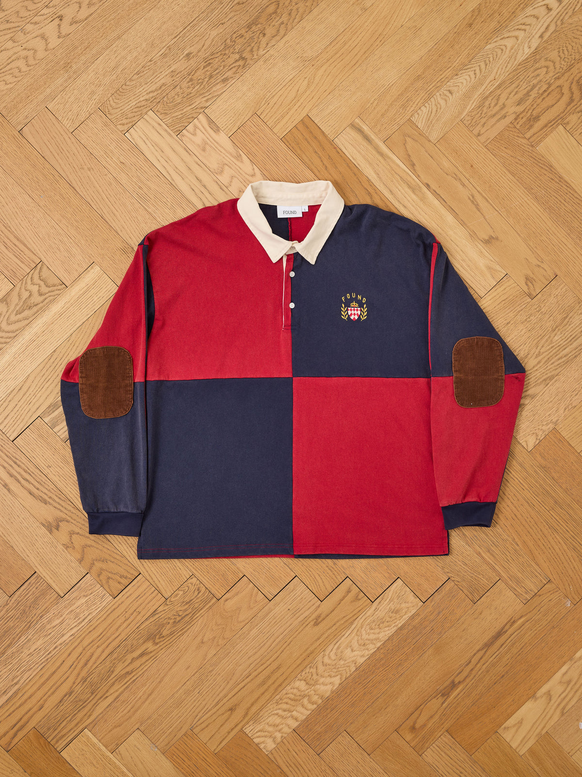 The FOUND Crestfield Rugby is a unisex long-sleeve shirt with bold red and navy color blocks, a crisp white collar, brown elbow patches, and a small crest logo on the chest, shown laid flat on a wooden parquet floor.