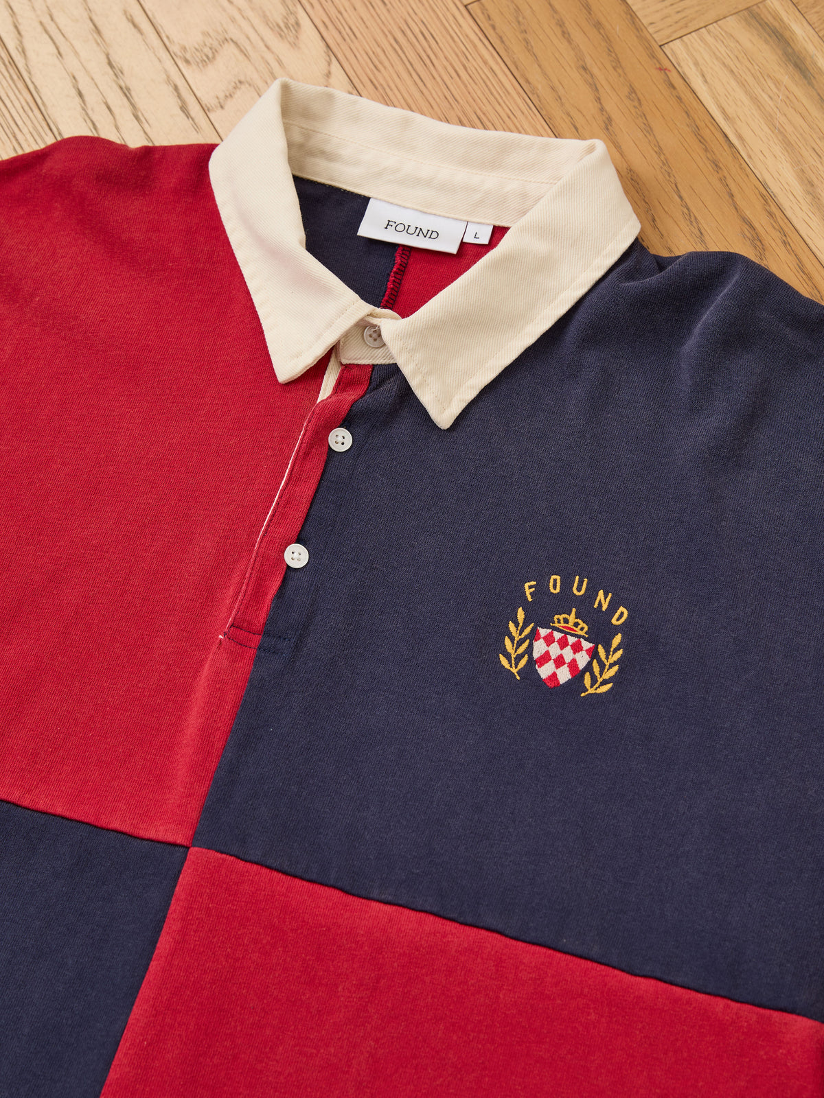 The Crestfield Rugby by FOUND is a unisex rugby shirt featuring color-blocked red and navy panels, a beige collar, and an embroidered “FOUND” crest on the chest, displayed flat on a wooden floor.
