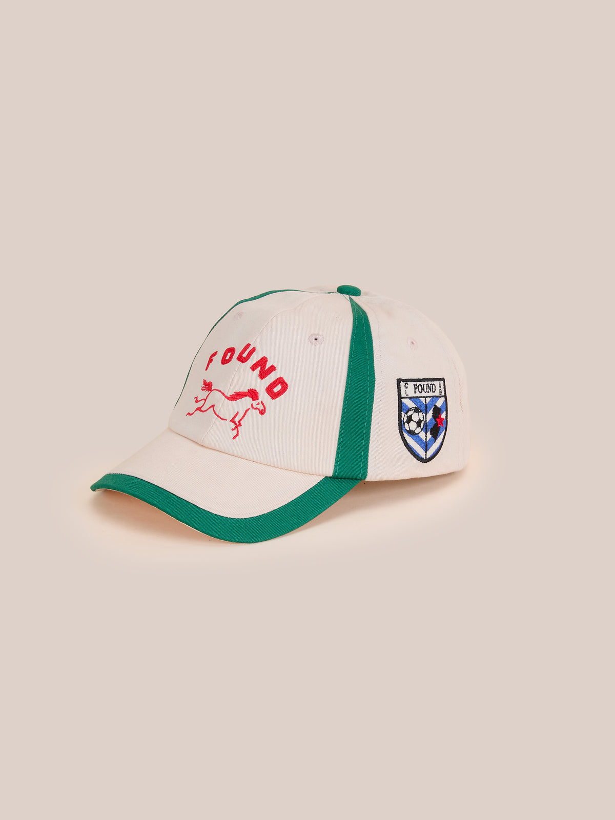 The Crestline Racing Cap by Found features beige fabric, green trim, red “FOUND” text, a running dog on the front, and a shield patch for a vintage equestrian look.