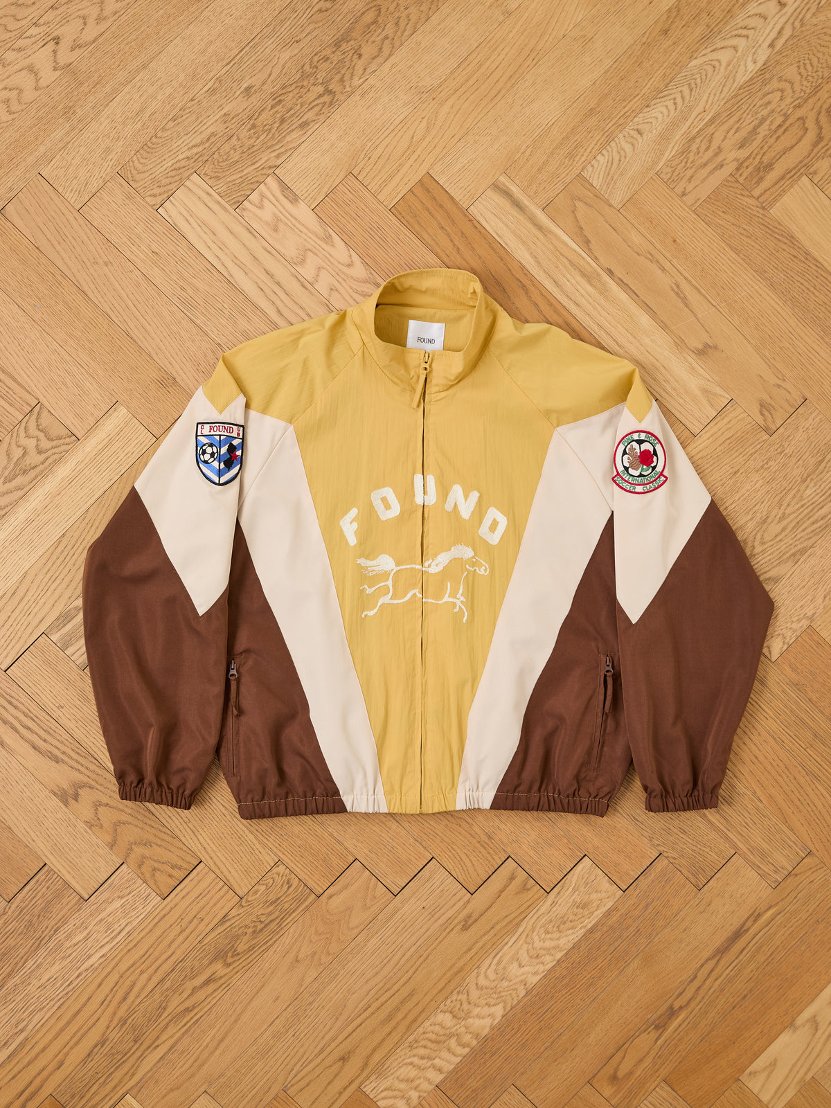 The Crestline Sport Zip Jacket by FOUND is a color-block shell with “FOUND” text, running dog graphic, and sleeve patches, shown laid flat on a wooden floor.