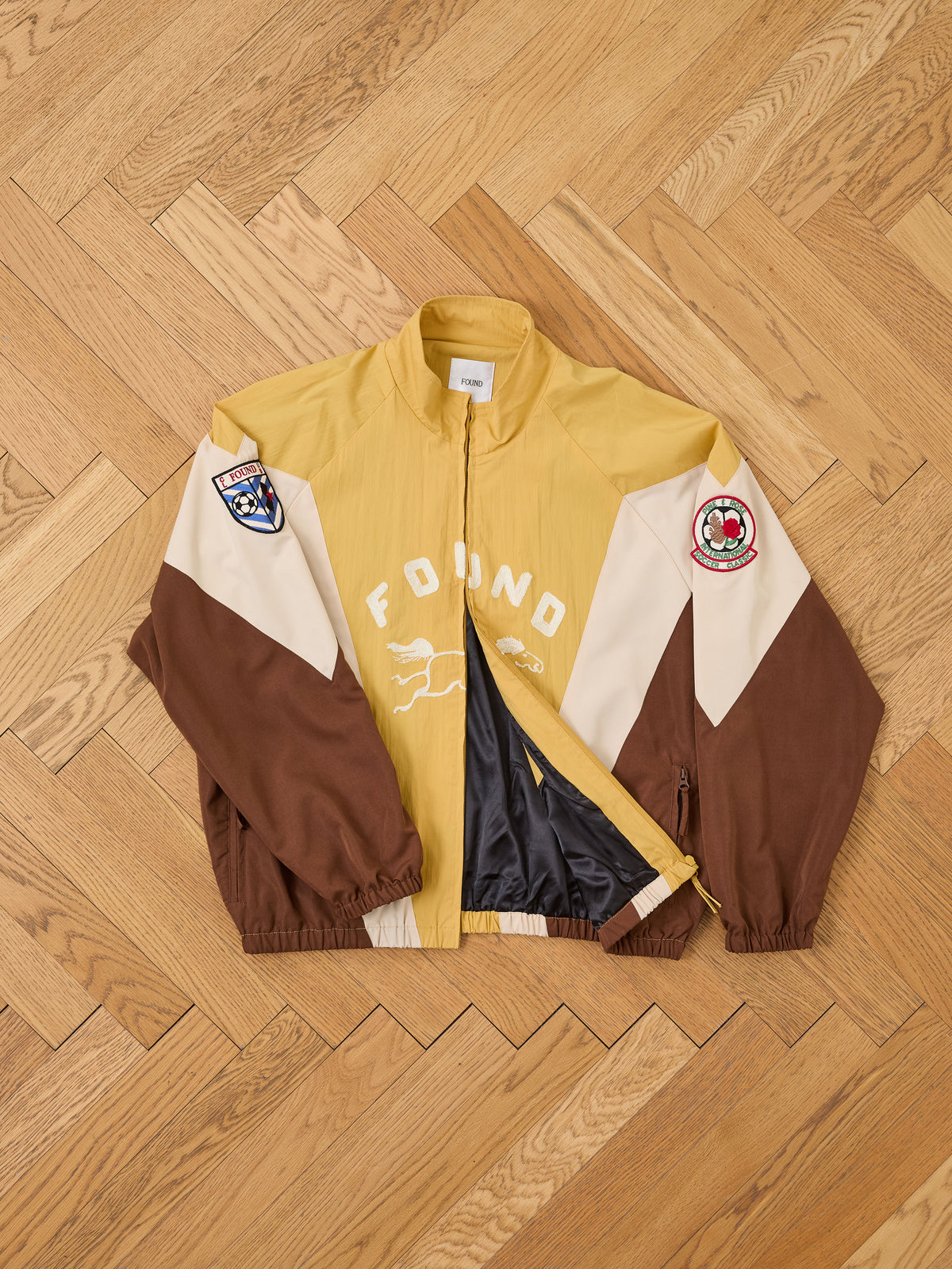 A yellow and brown unisex Crestline Sport Zip Jacket by FOUND, featuring embroidered patches, a white “FOUND” logo, and animal graphic, laid on a wooden parquet floor.