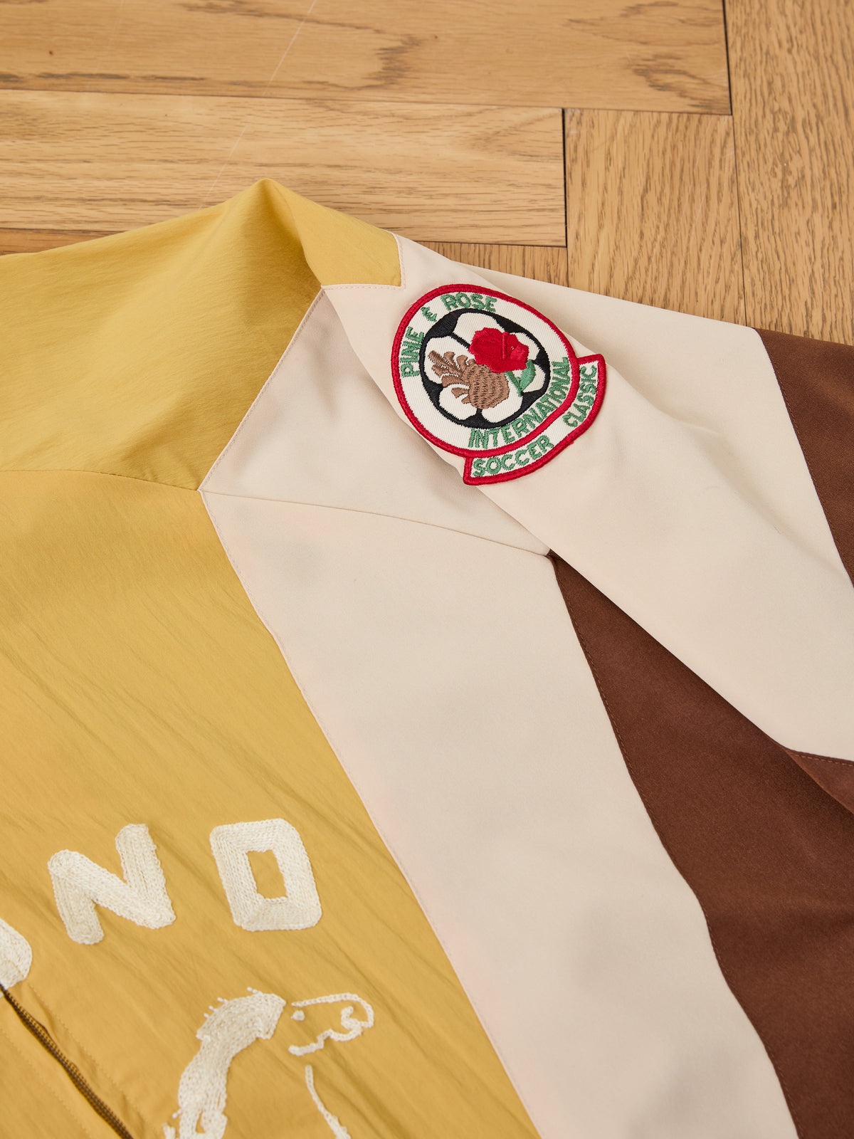 A close-up of a yellow, cream, and brown Crestline Sport Zip Jacket by FOUND with an embroidered rose patch reading “Mister Diamond,” laid on a wooden floor.
