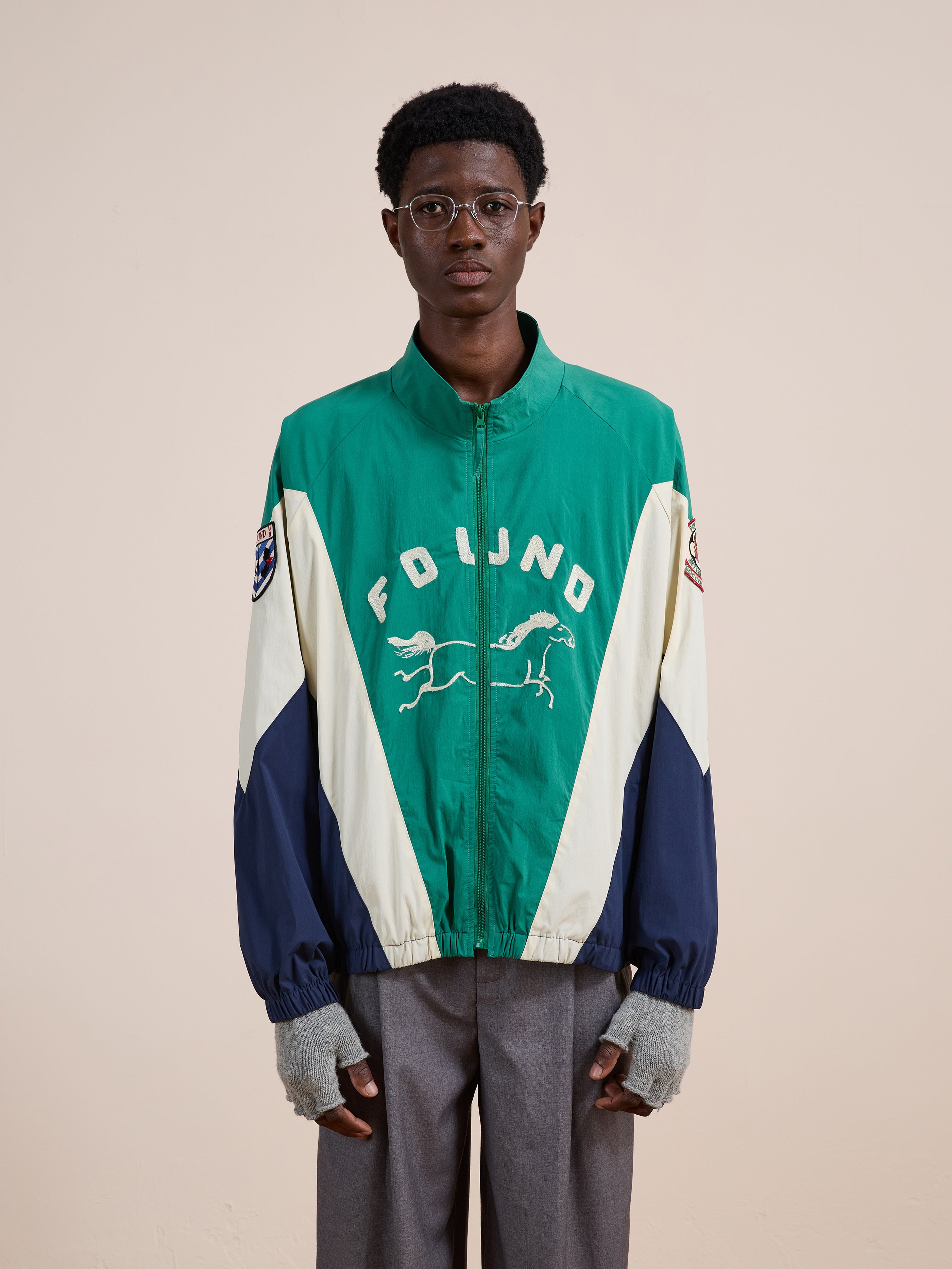 A person faces forward, wearing glasses, gray trousers, fingerless gloves, and the FOUND Crestline Sport Zip Jacket in green and white with a horse graphic, standing against a plain background.