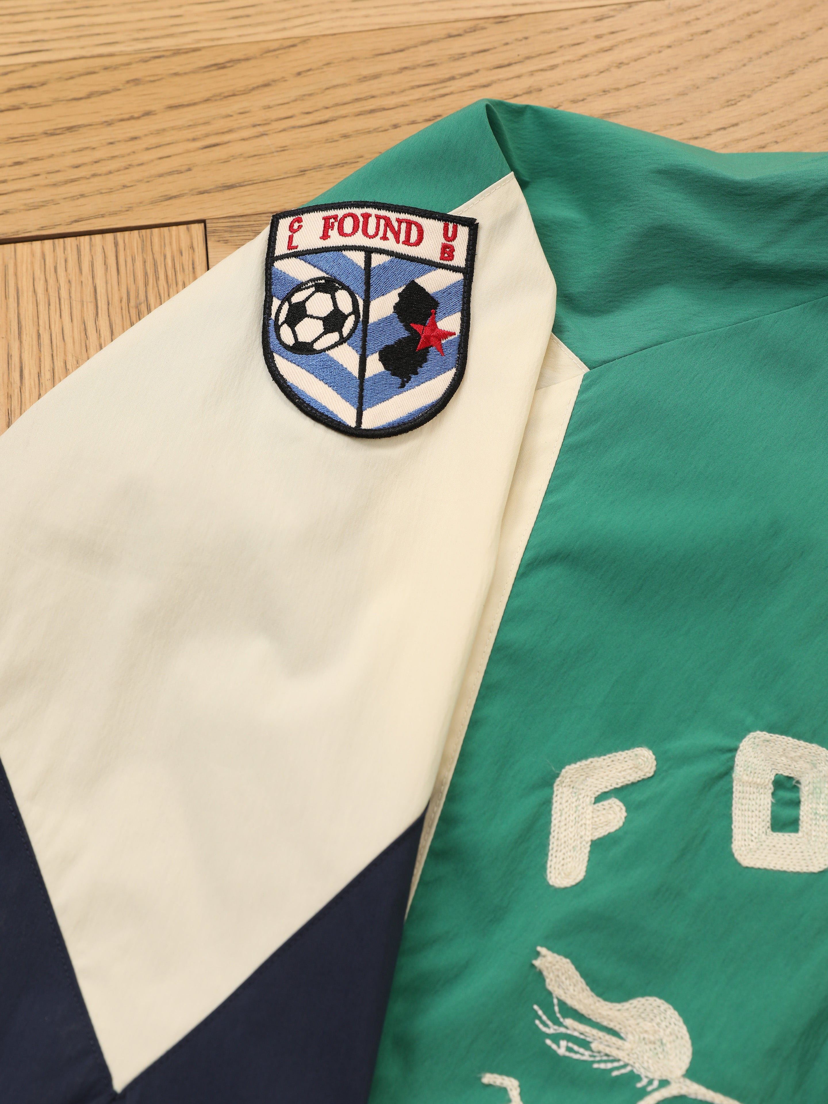 The FOUND Crestline Sport Zip Jacket is a vintage green and white windbreaker with a sleeve patch featuring a soccer ball, star, and "FOUND." Lightweight and sporty, it's perfect for casual or athletic wear.