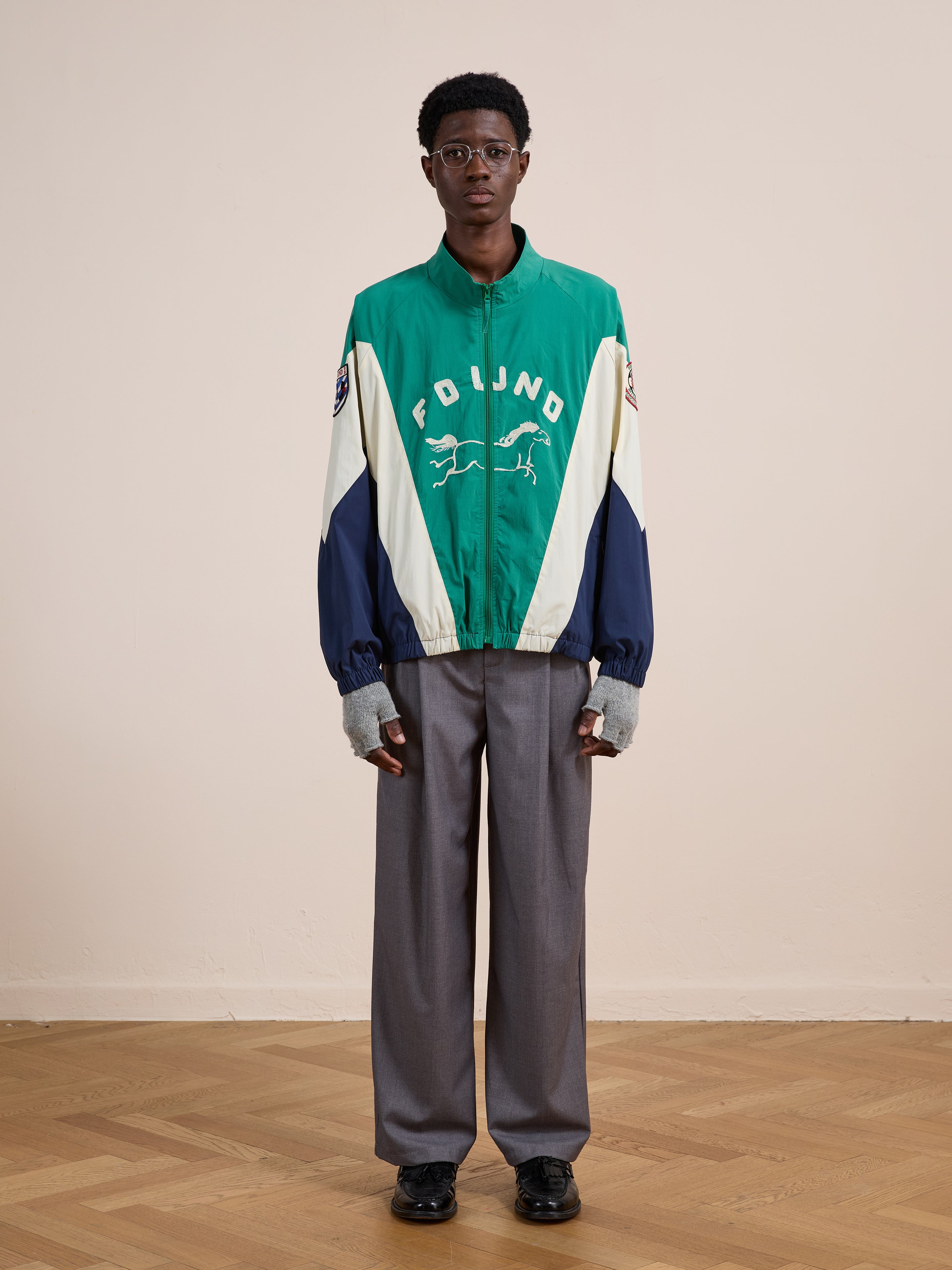 Indoors against a beige wall and wooden floor, a person wears a green and white FOUND Crestline Sport Zip Jacket, gray wide-leg trousers, dark shoes, and eyeglasses—channeling classic vintage sportswear style.