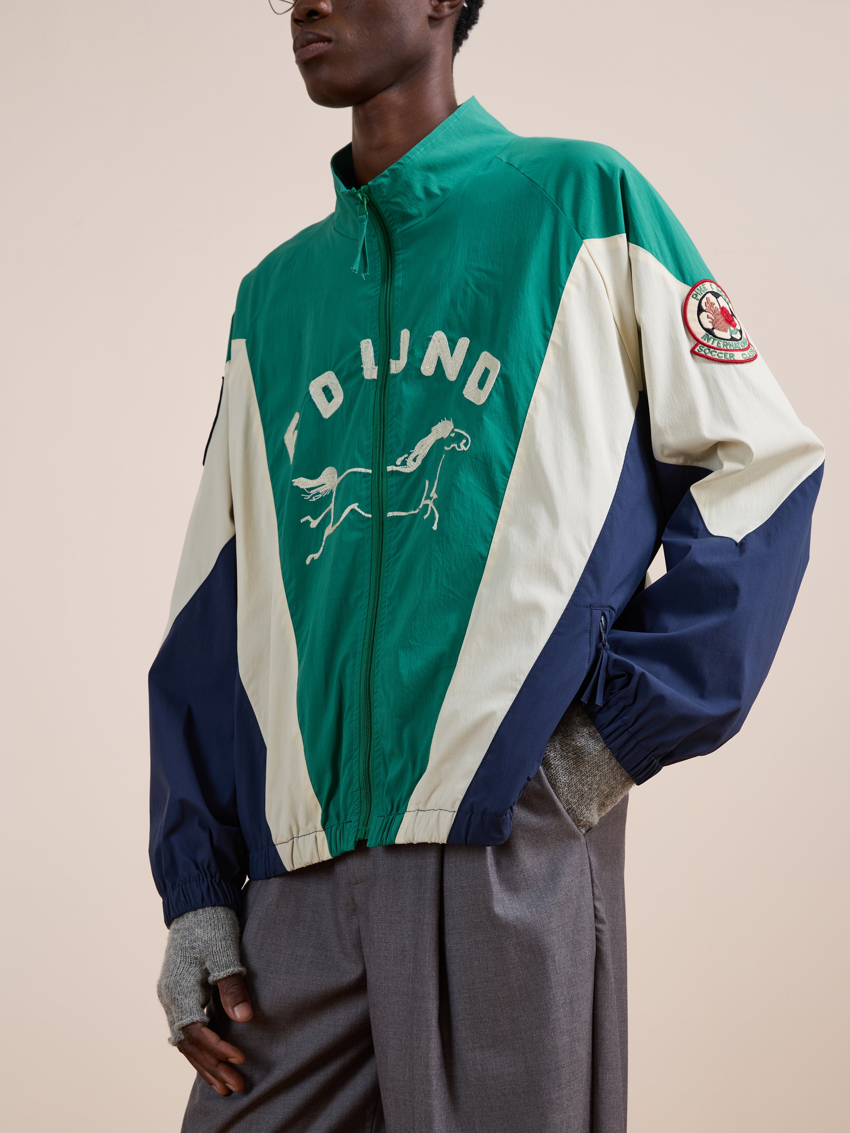 Someone in a green, white, and navy FOUND Crestline Sport Zip Jacket featuring "FOUND" and a running dog graphic on the front, paired with gray pants and gray fingerless gloves for a vintage sportswear-inspired look.