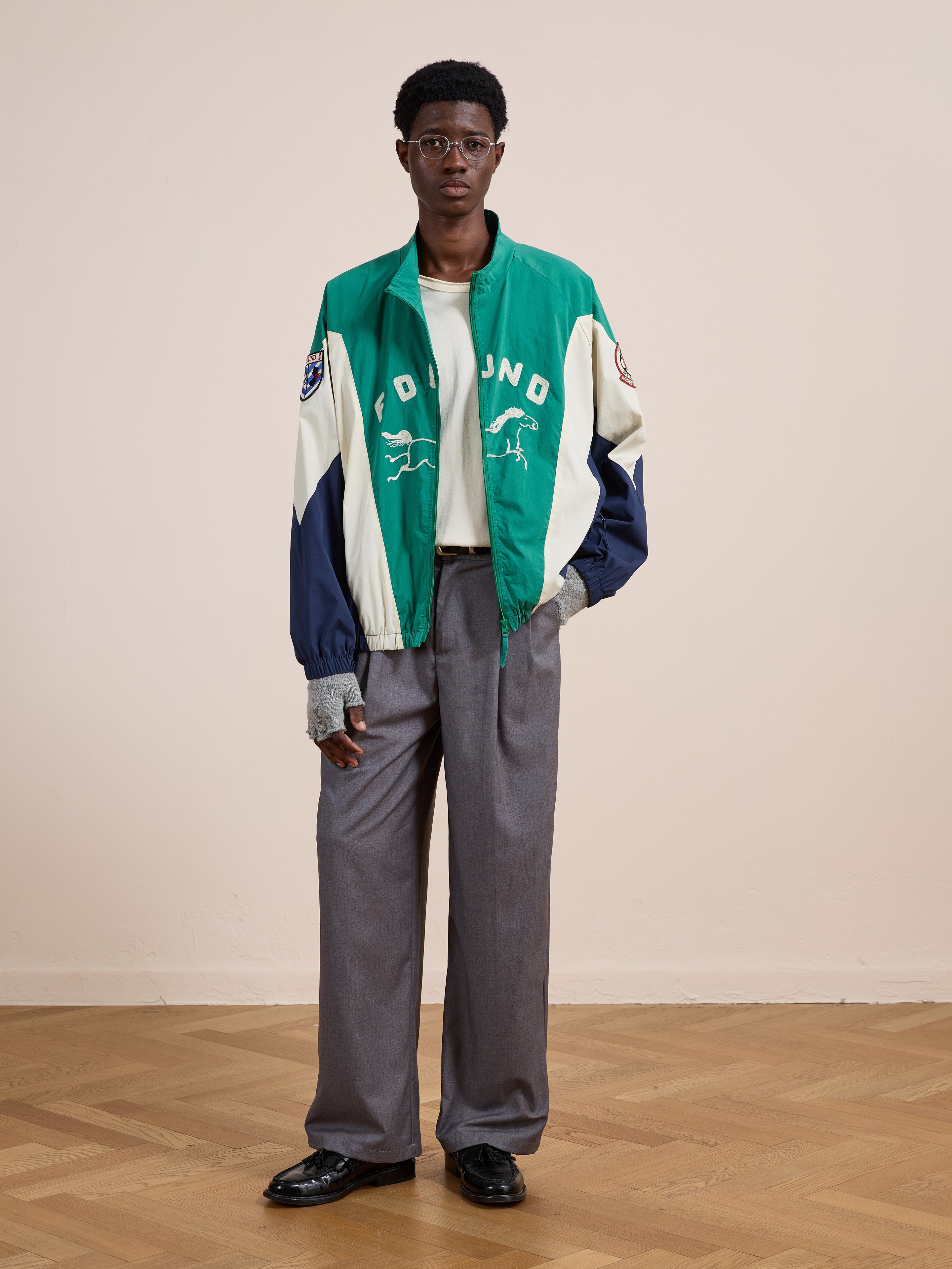 Indoors on a wooden floor, someone wears the FOUND Crestline Sport Zip Jacket in green and cream with text and graphics, paired with gray wide-leg trousers, a white shirt, and black shoes—a modern vintage sportswear style.